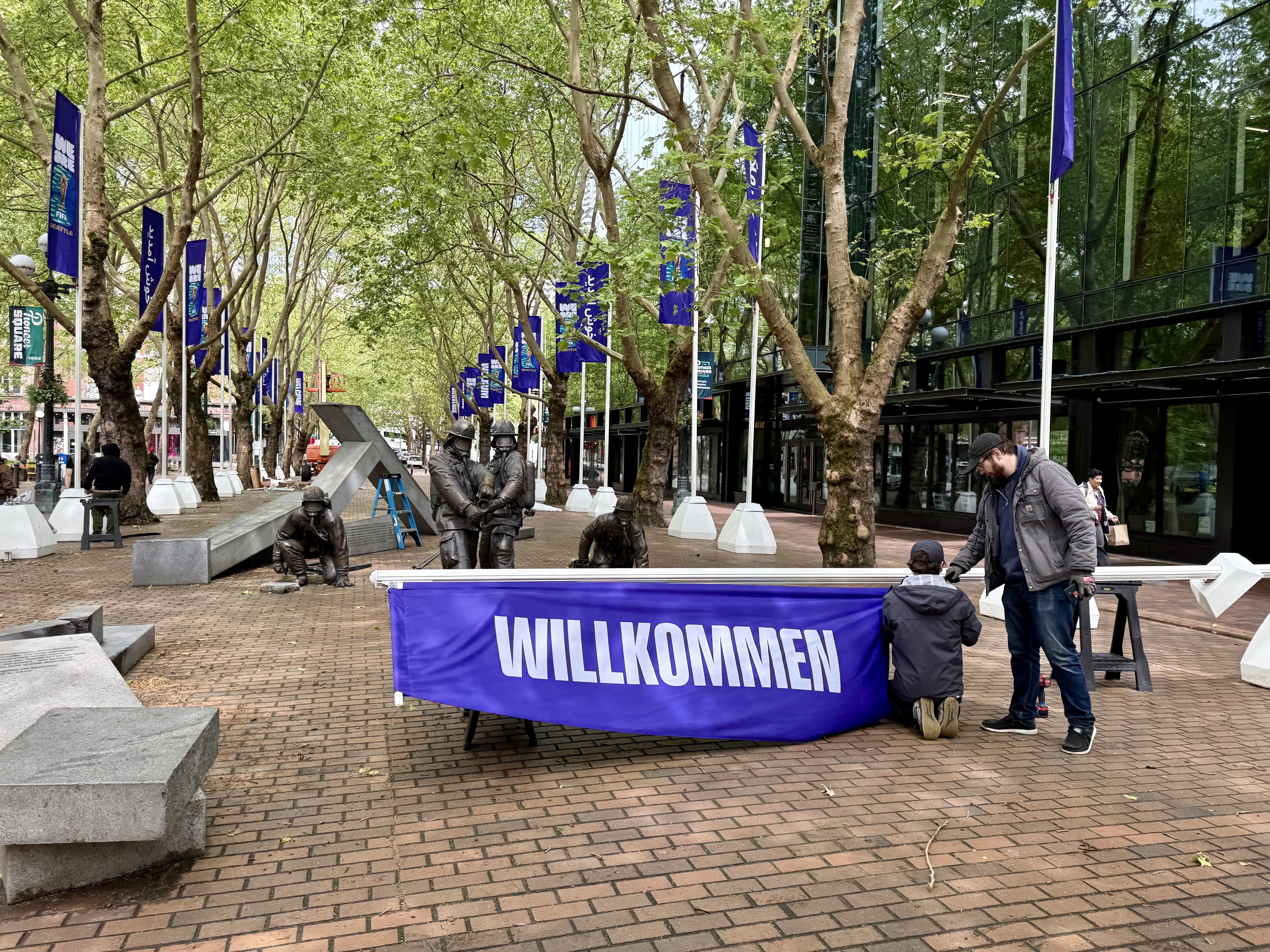 Tree-lined city plaza with purple banners along a glass-front building. A bronze group sculpture sits in the foreground as workers set up equipment and a ladder nearby.