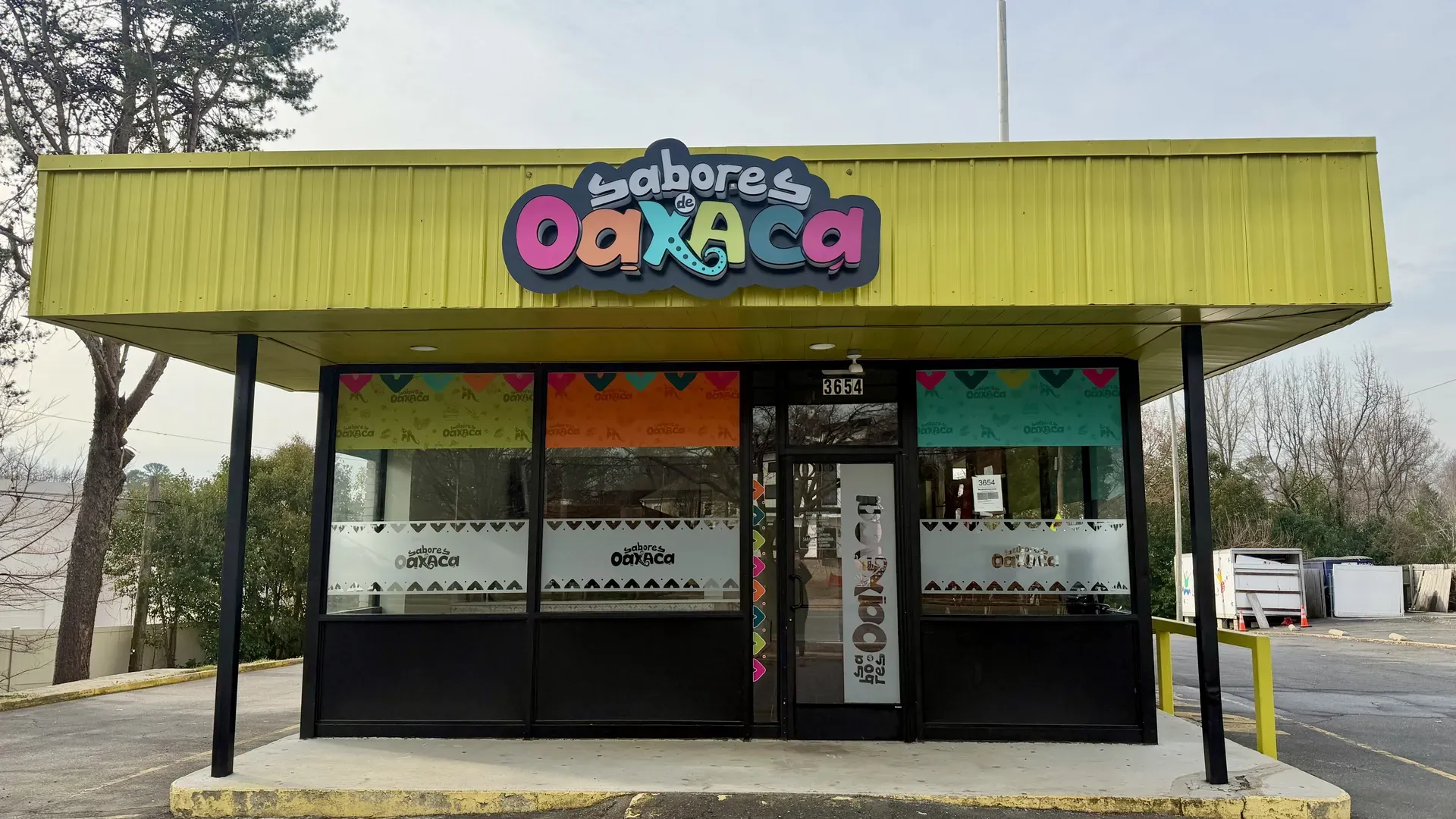 Bright yellow storefront with a large, colorful sign reading "Sabores de Oaxaca" above the entrance. Black-framed windows with decorative panels; address 3654 over the door; blue sky and trees nearby.