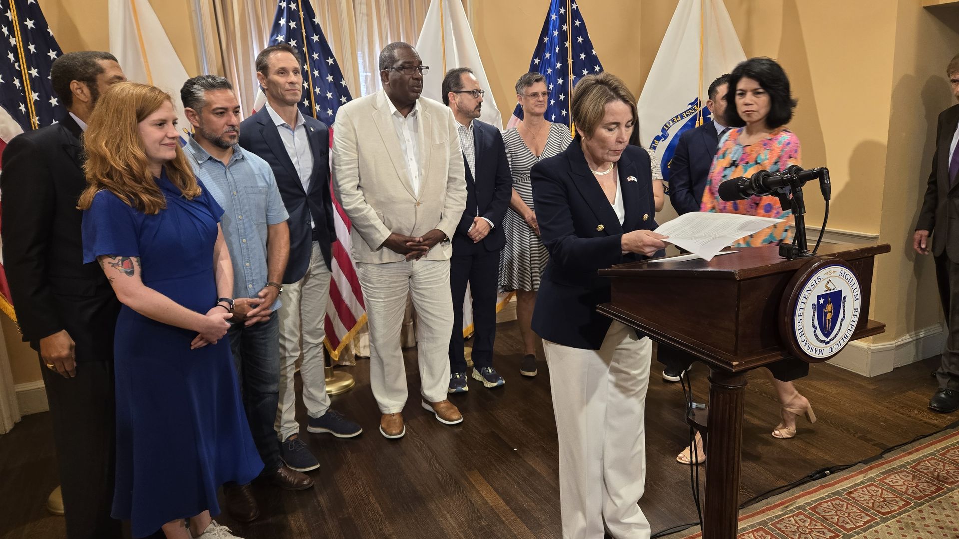 A woman in a navy blazer and white pants reads from papers at a podium with the Massachusetts seal, surrounded by a diverse group of people standing in front of American and state flags indoors.