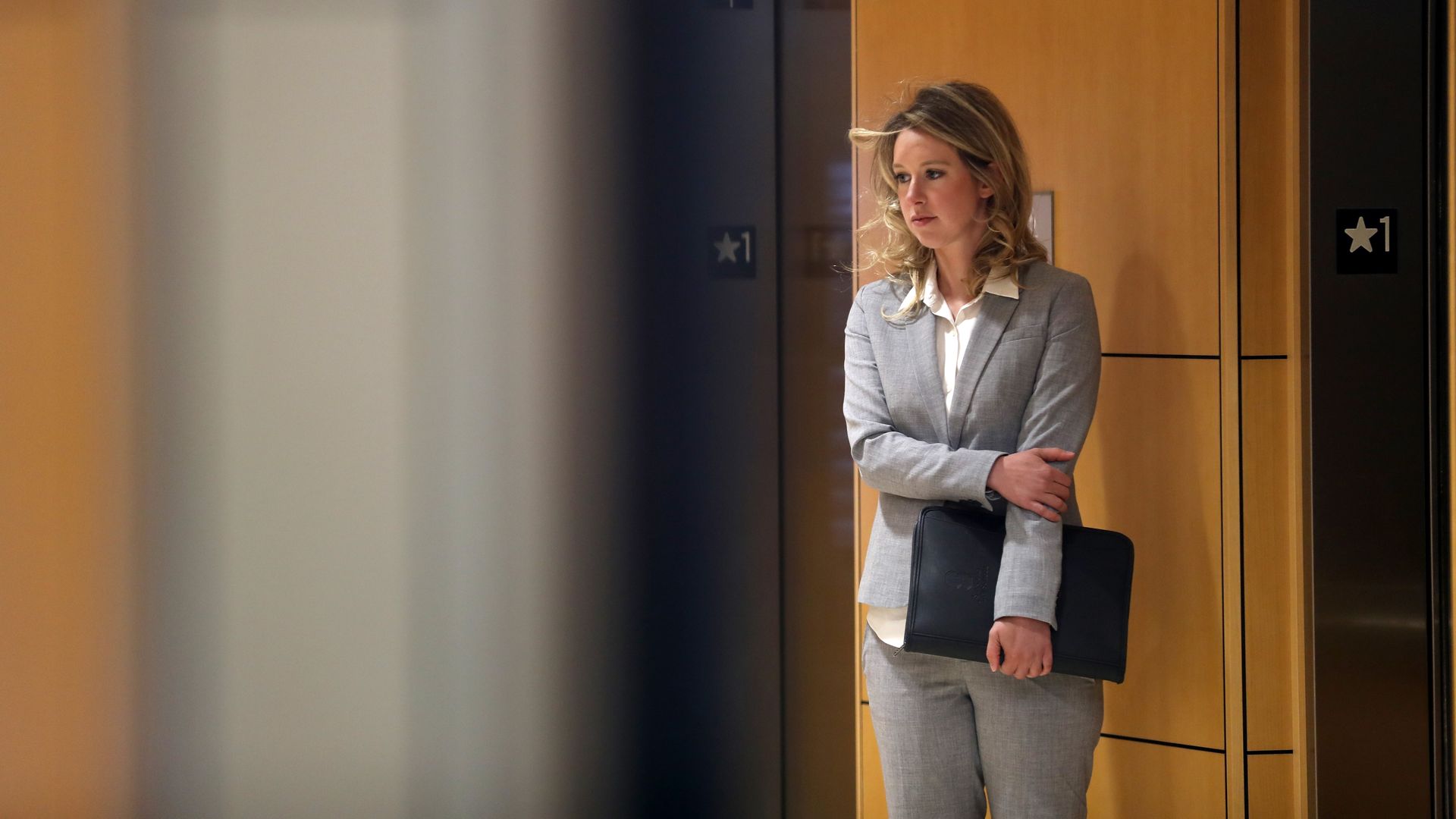 In this image, Holmes stands holding an arm across her stomach while waiting for an elevator.