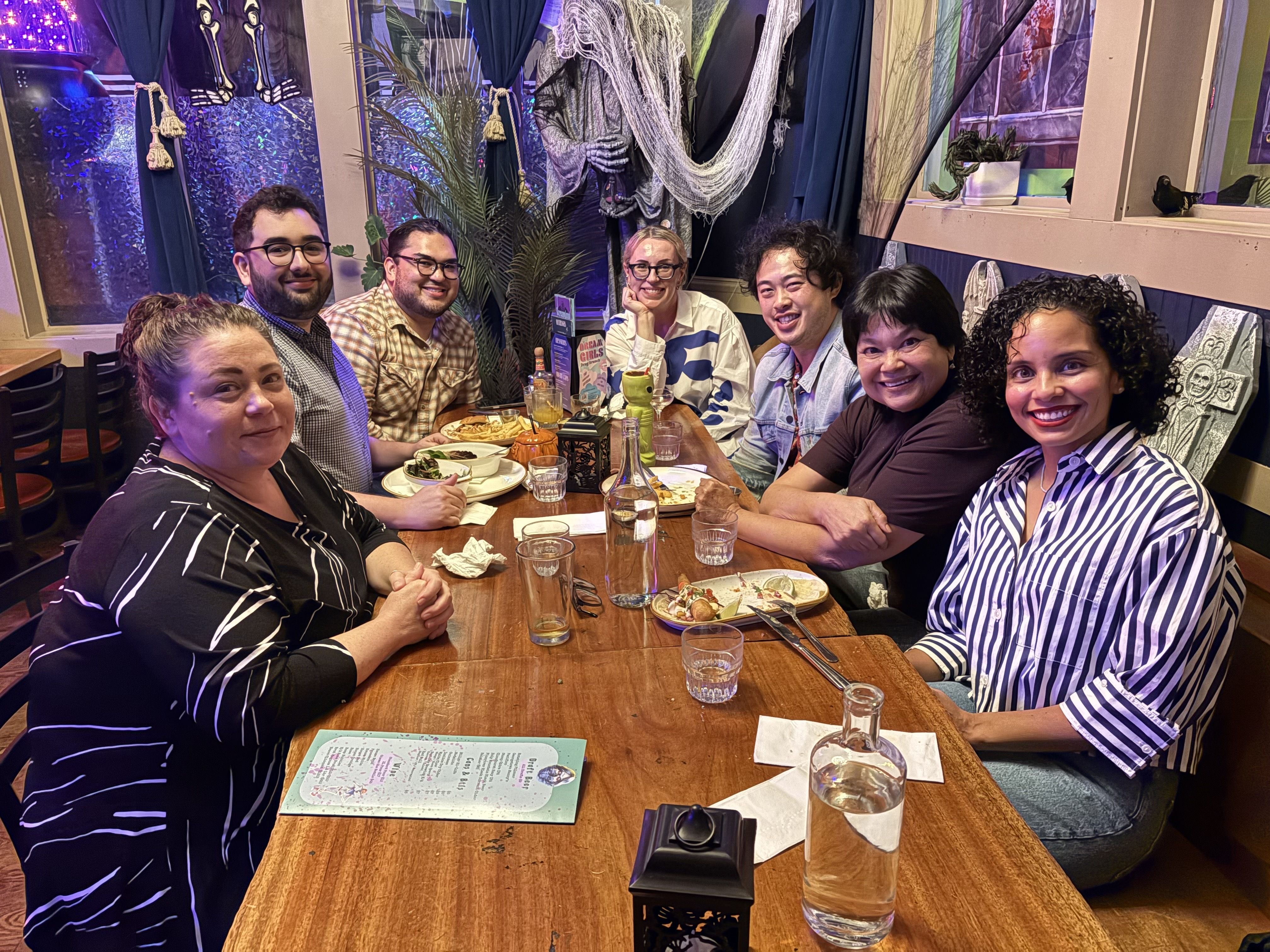 Seven people sit around a long wooden table in a colorful, tropical-style restaurant, smiling at the camera. Plates and drinks are on the table; vibrant curtains and potted plants decorate the background.