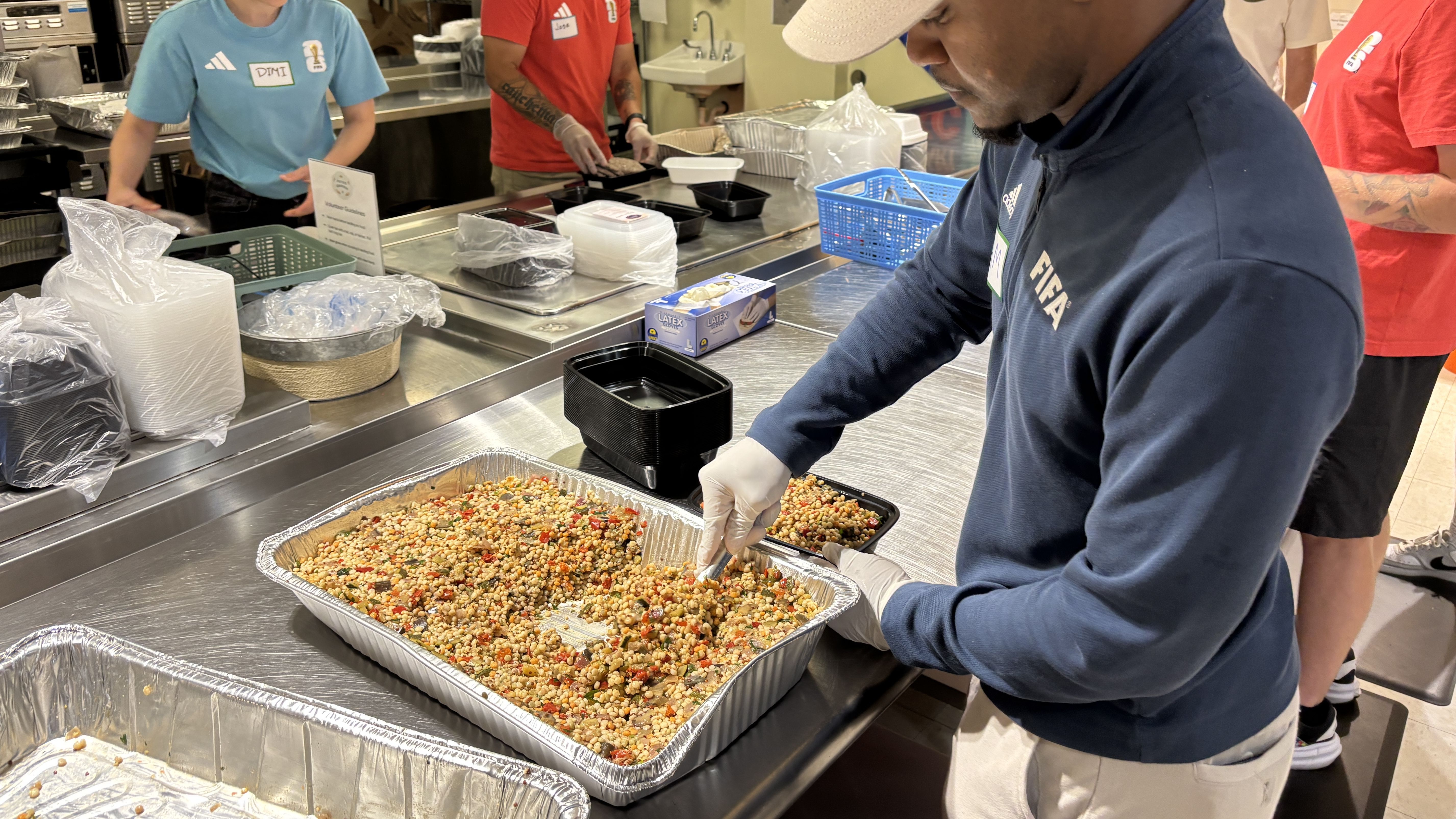 Several volunteers work behind a cafeteria line: a man in a dark blue jacket that says "FIFA" and gloves scoops a colorful mixed bean and corn dish from a large aluminum tray onto a smaller tray, as others prepare food in the background.