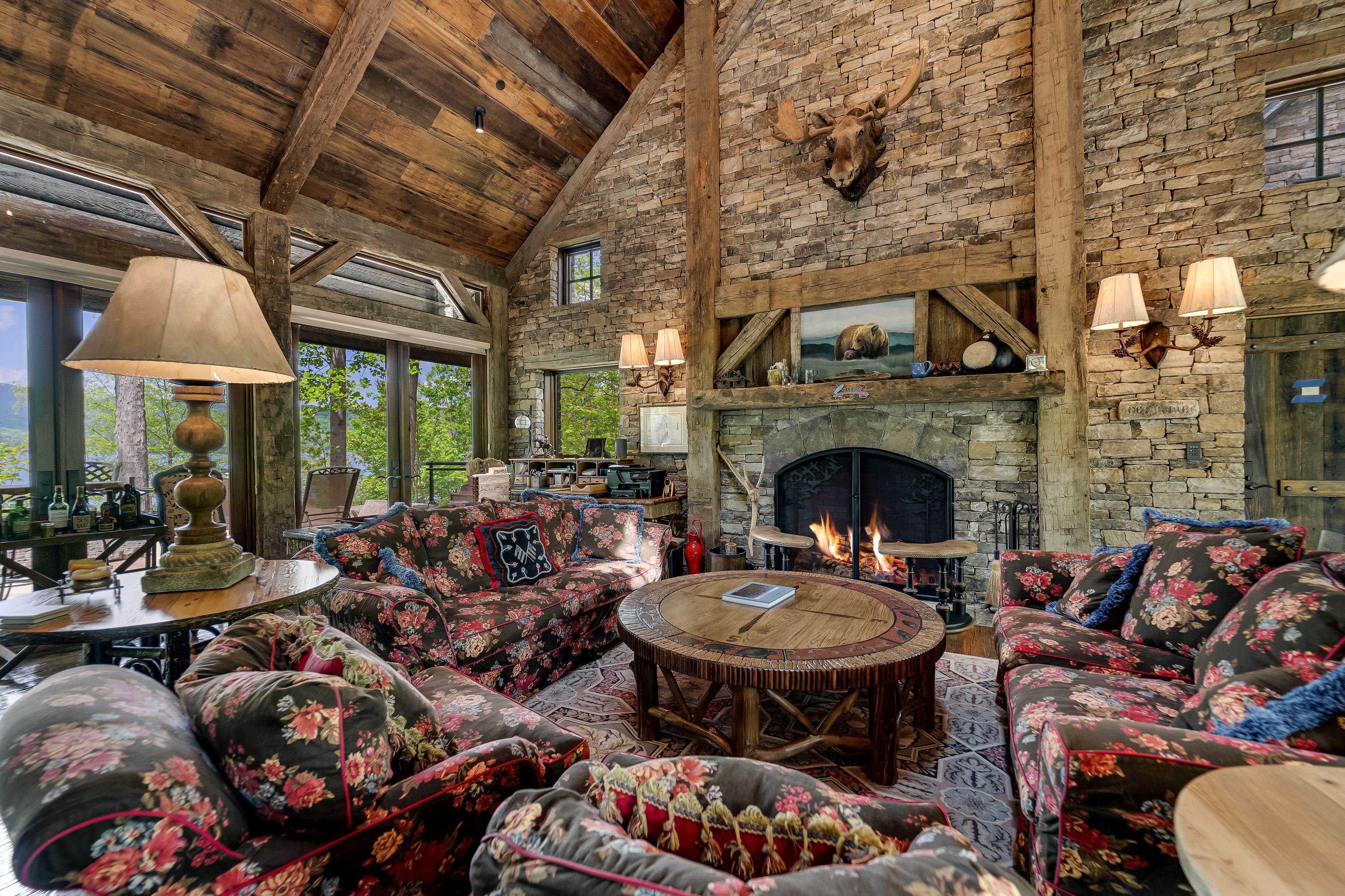 Cozy rustic living room with floral sofas, wooden round table, stone fireplace with fire, mounted moose head, wood beams, stone walls, and large windows showing greenery outside.