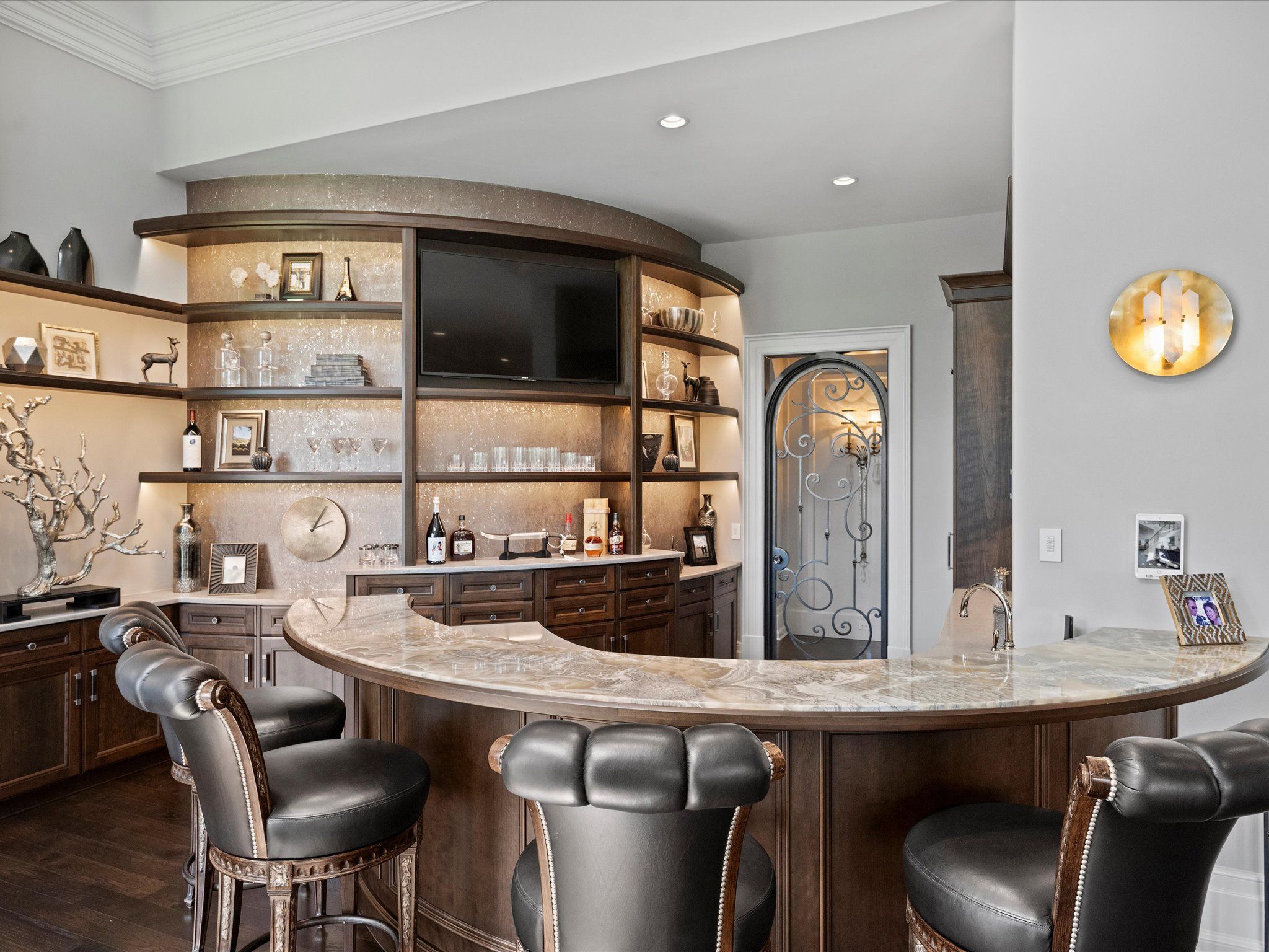 Curved bar with marble countertop and dark wood base, surrounded by black leather cushioned bar stools. Behind, illuminated shelves with decorative items, bottles, and a TV screen.