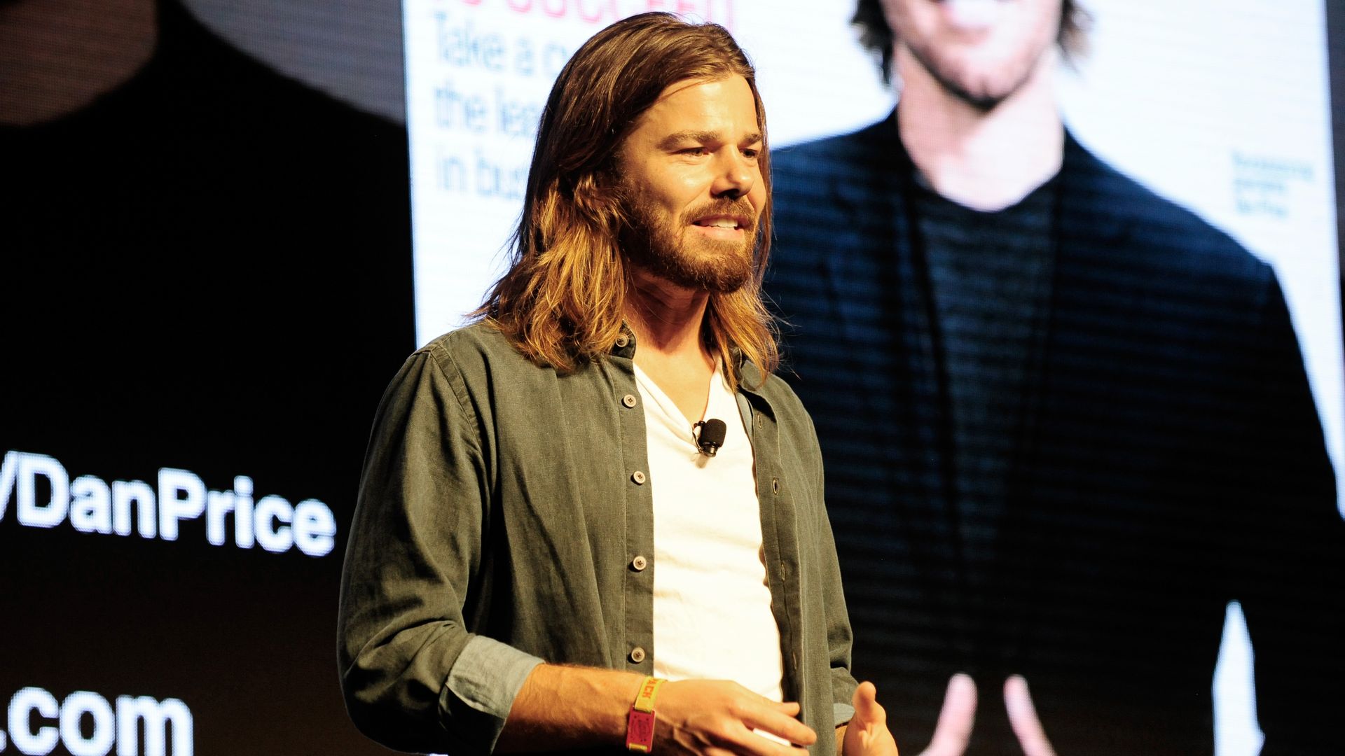 A man in a light green outershirt with rolled up sleeves over a white shirt, and shoulder length brown hair, on a state with a magazine cover projected behind him.