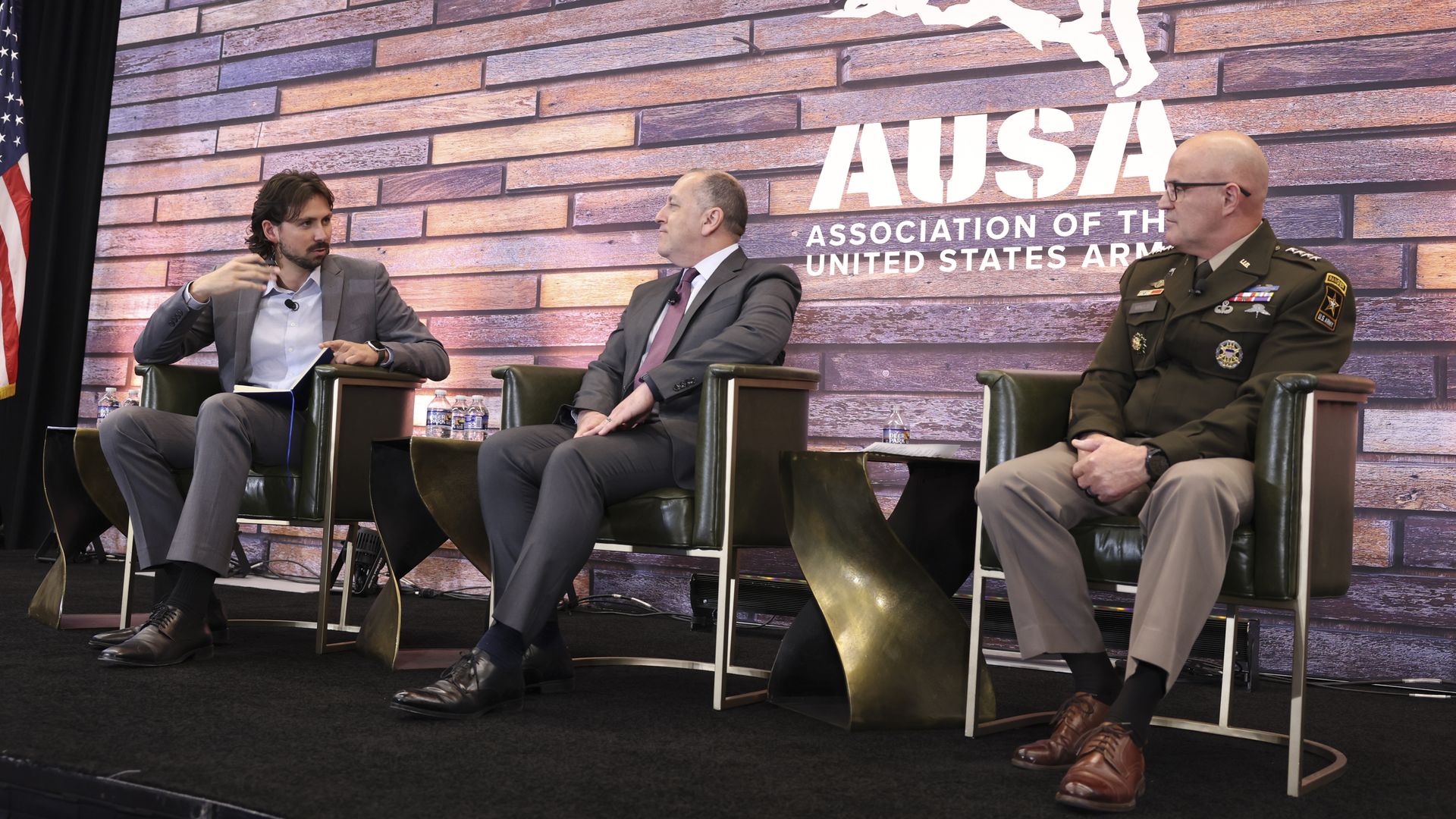 Three men sit on a stage in green chairs. The carpet is black. A brick wall is behind them with text that says, 