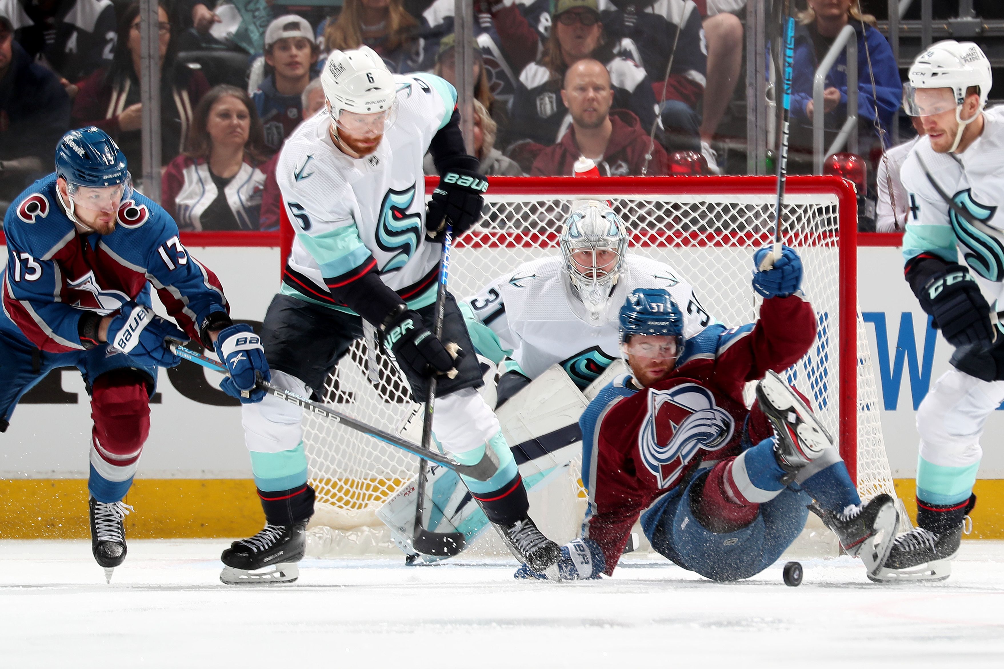 Five hockey players are shown with three in the forefront, one of them on the ground, with a goal in background.