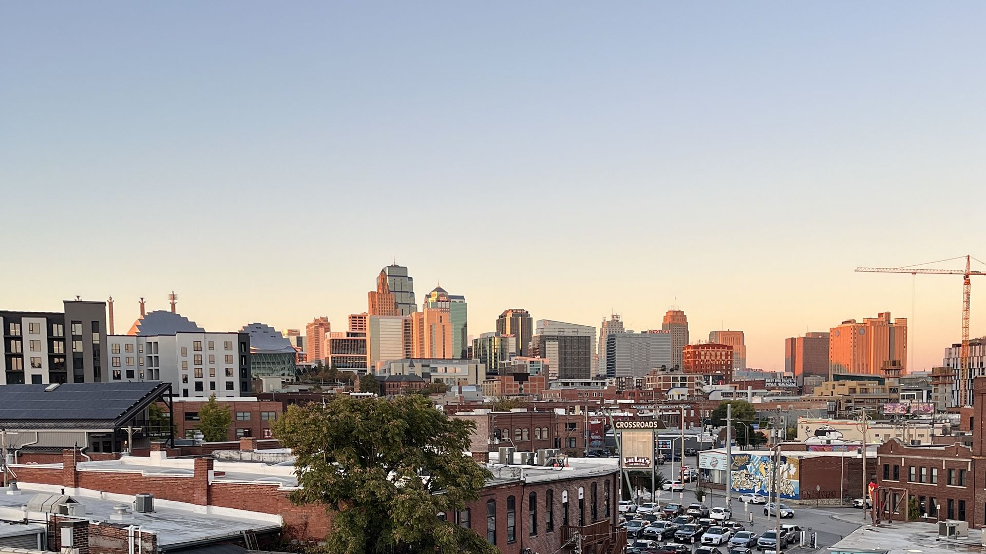 Sunset Kansas City skyline with a blue-to-peach gradient sky. Foreground of Crossroads brick rooftops and a tree, midrise buildings across the horizon, and a construction crane on the right.