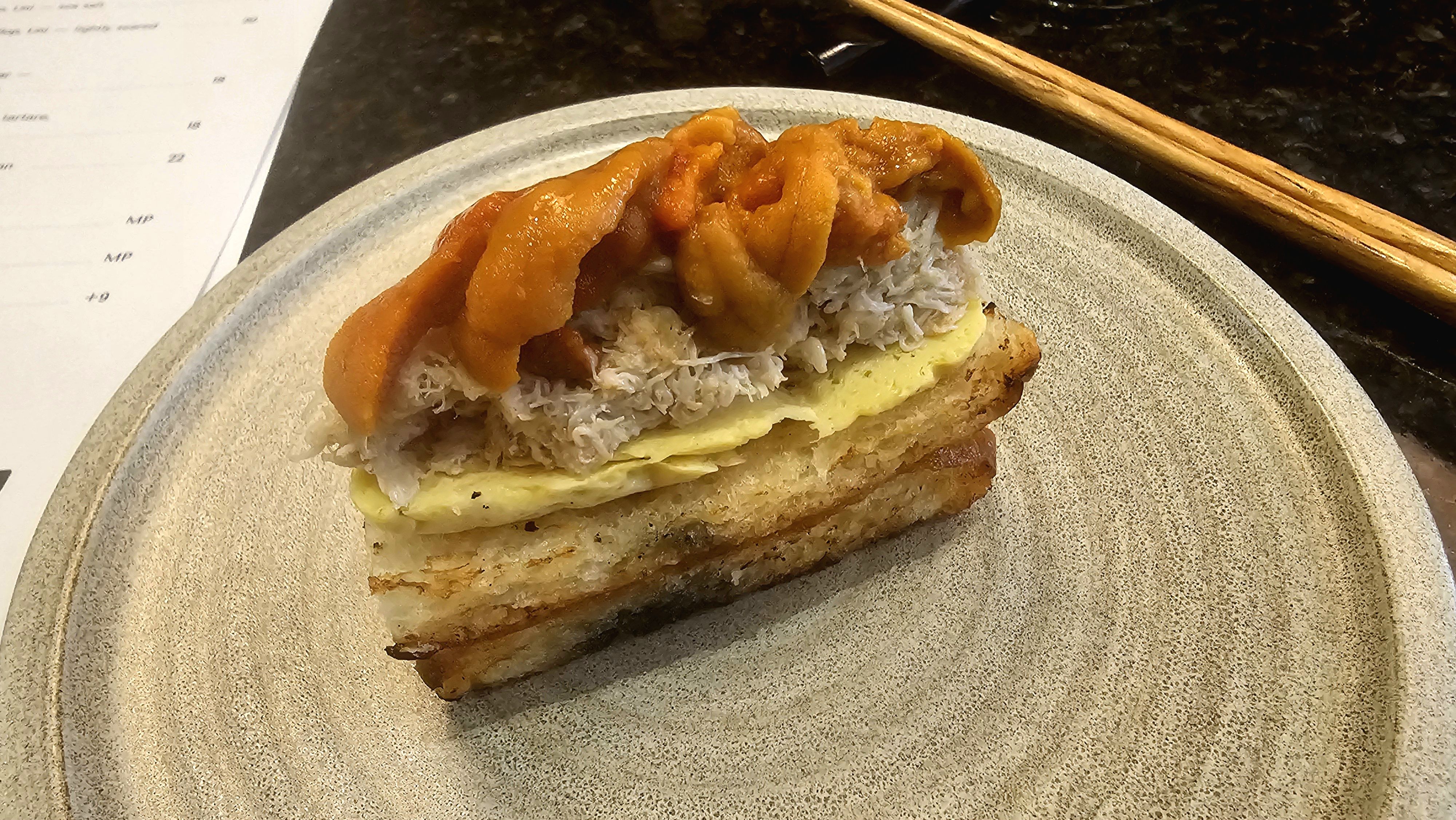 Open-faced toast with a yellow egg layer, white shredded crab, and bright orange uni (sea urchin) on top, on a speckled plate with chopsticks nearby.