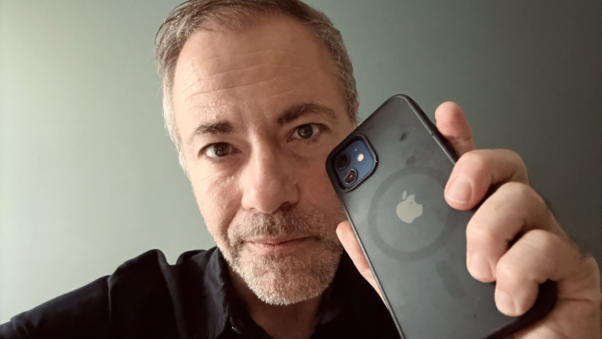 Close-up of a middle-aged man with gray stubble, holding a blue-gray iPhone to his cheek to take a selfie. He wears a dark shirt, against a pale green wall.
