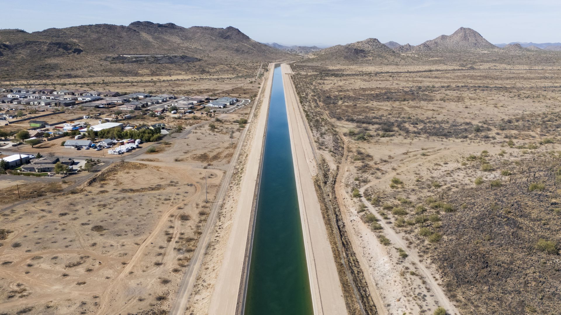 A canal carries water through a brown desert landscape into a mountainous area, with houses on one side and open scrubland on the other. 