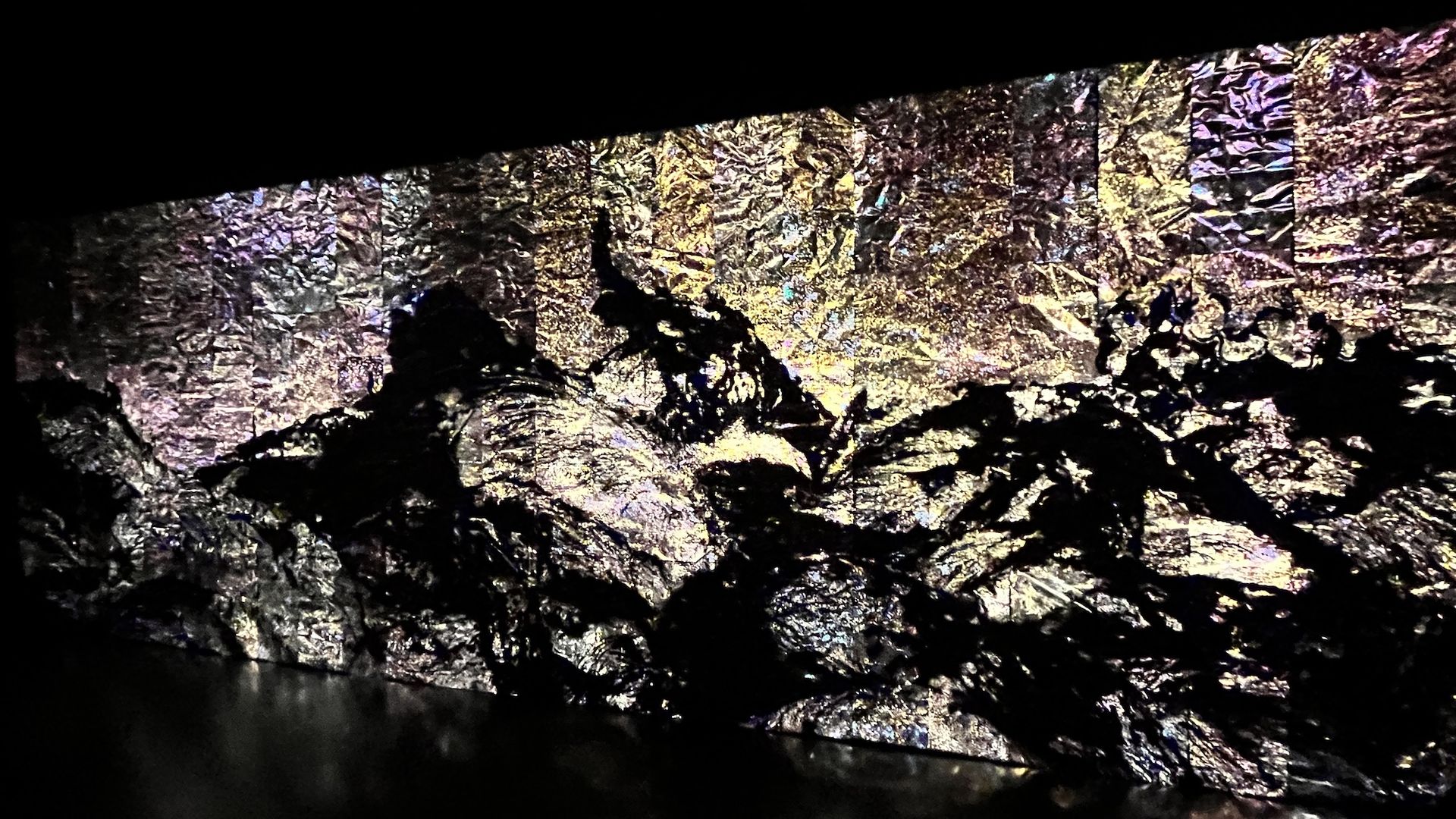 Crinkled metallic foil wall shimmering in gold, purple, and silver under dark lighting; a small silhouette stands near the center against a black upper background.