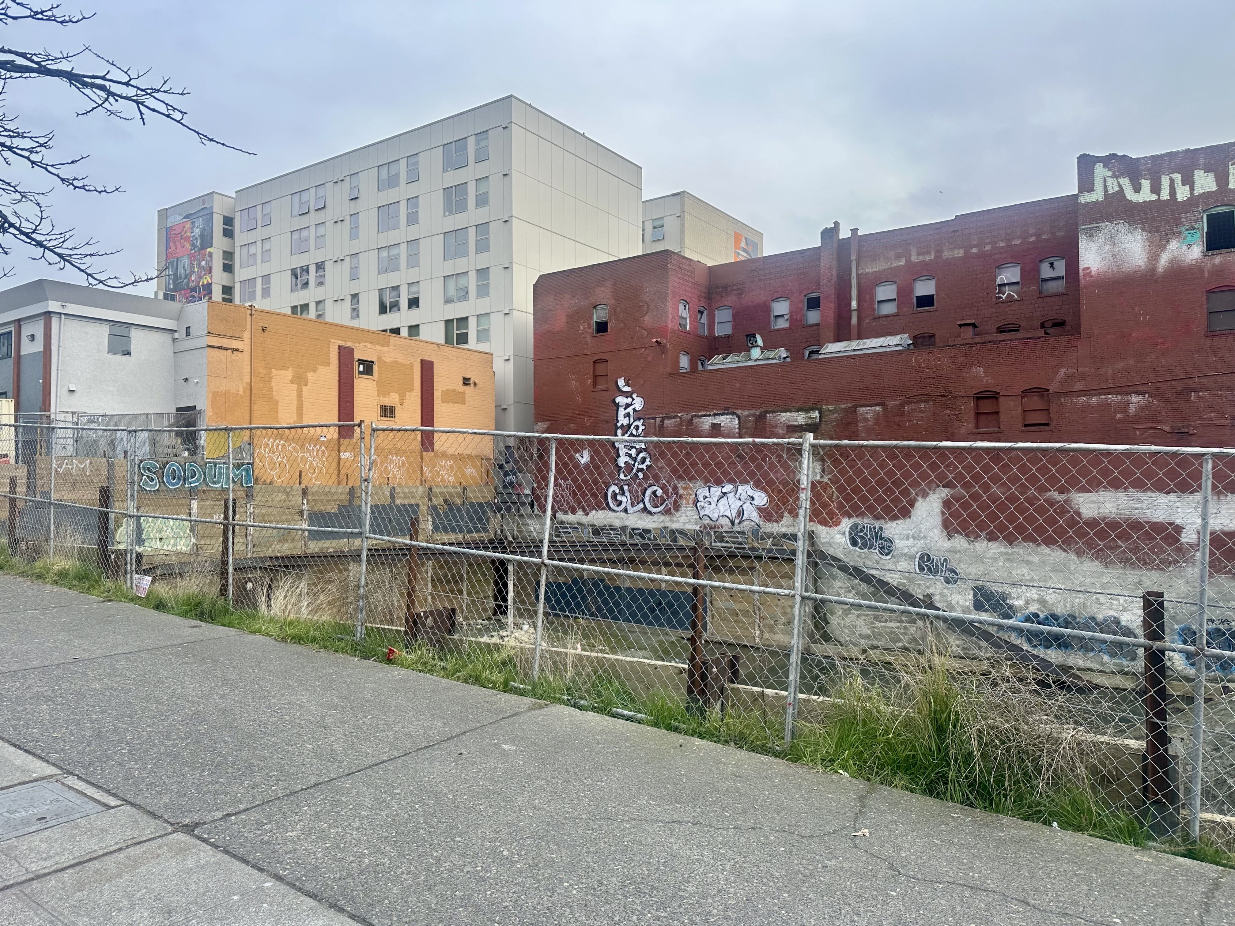 Urban scene with a chain-link fence in front of red and orange brick buildings covered in graffiti under a cloudy sky, with a modern white building in the background.