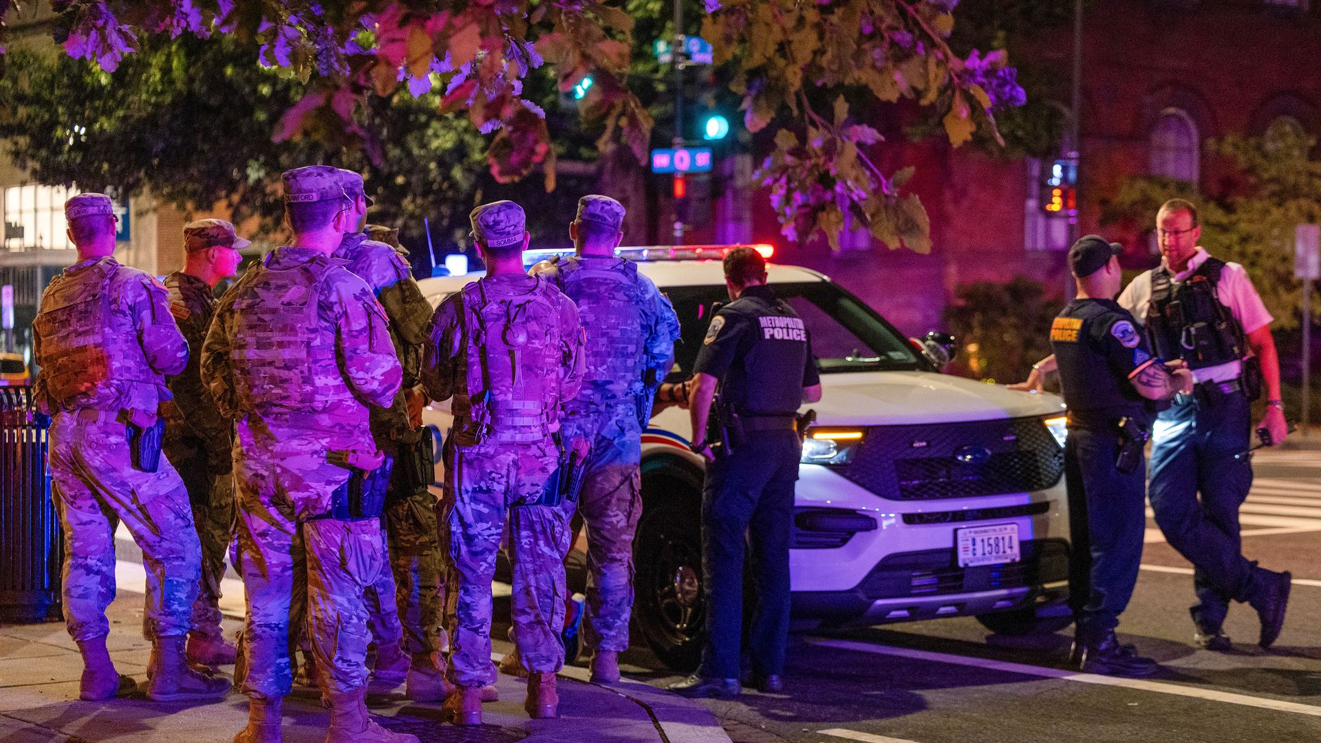 Group of uniformed military personnel and police officers gathered around a white police SUV at night, illuminated by streetlights and red-blue emergency lights.