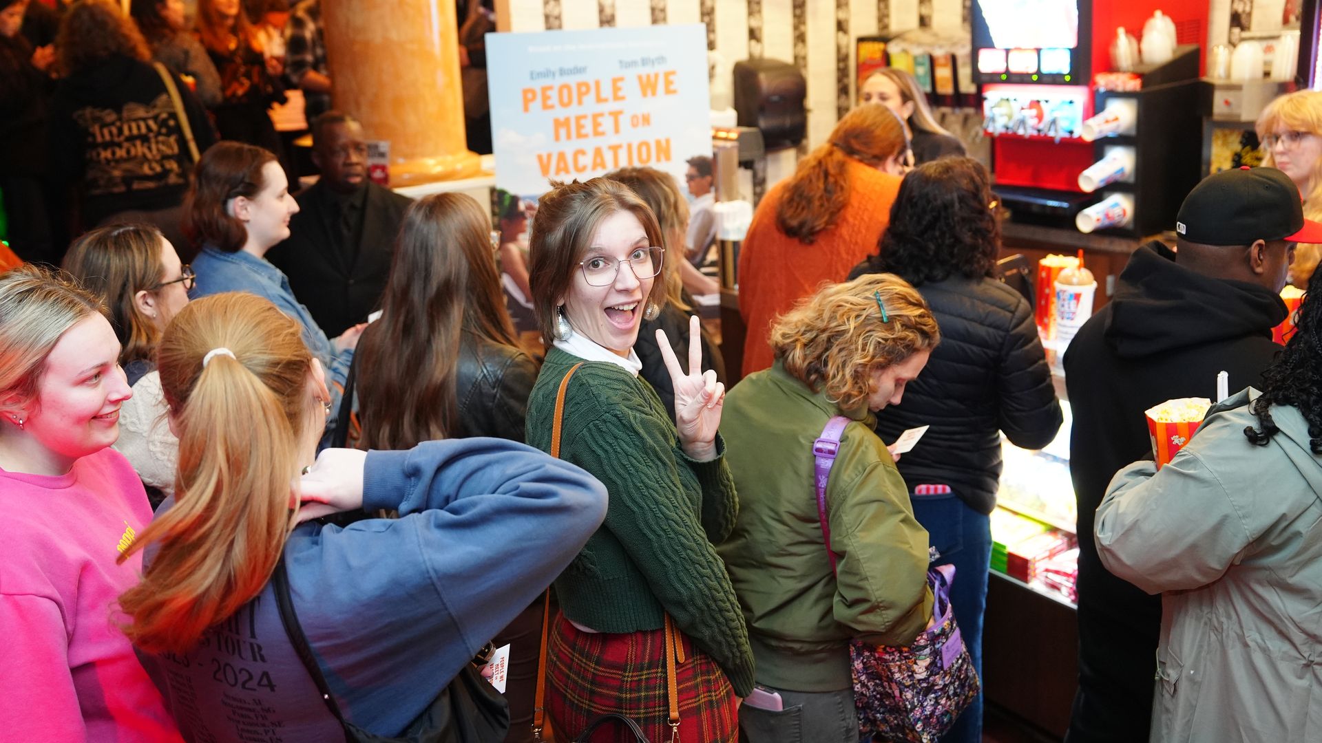 A woman flashes a peace sign over her shoulder as she stands in line for snacks at a movie theater. A movie poster for "People We Meet On Vacation" is spotted behind the crowd. 