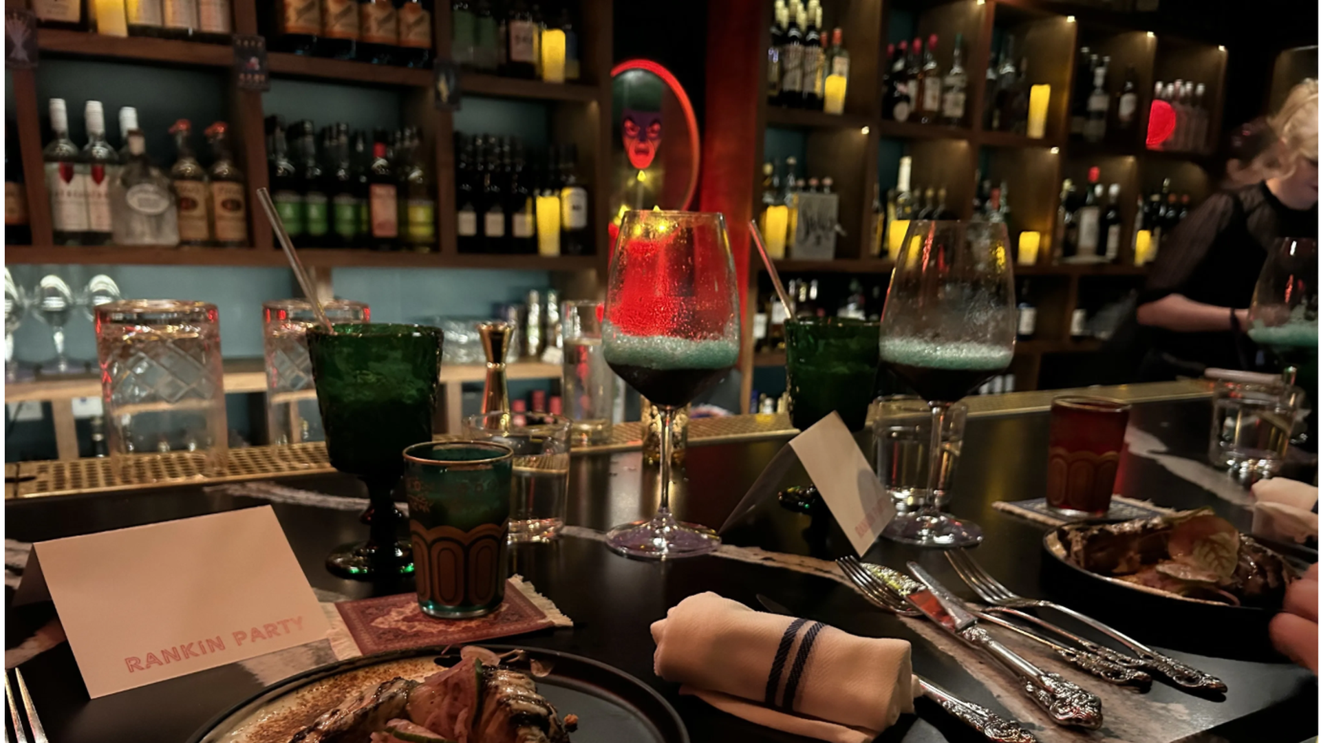 Bar setting with two plates of gourmet food, green and red glasses, silver cutlery, rolled napkins, and place cards labeled "Rankin Party" under warm ambient lighting.