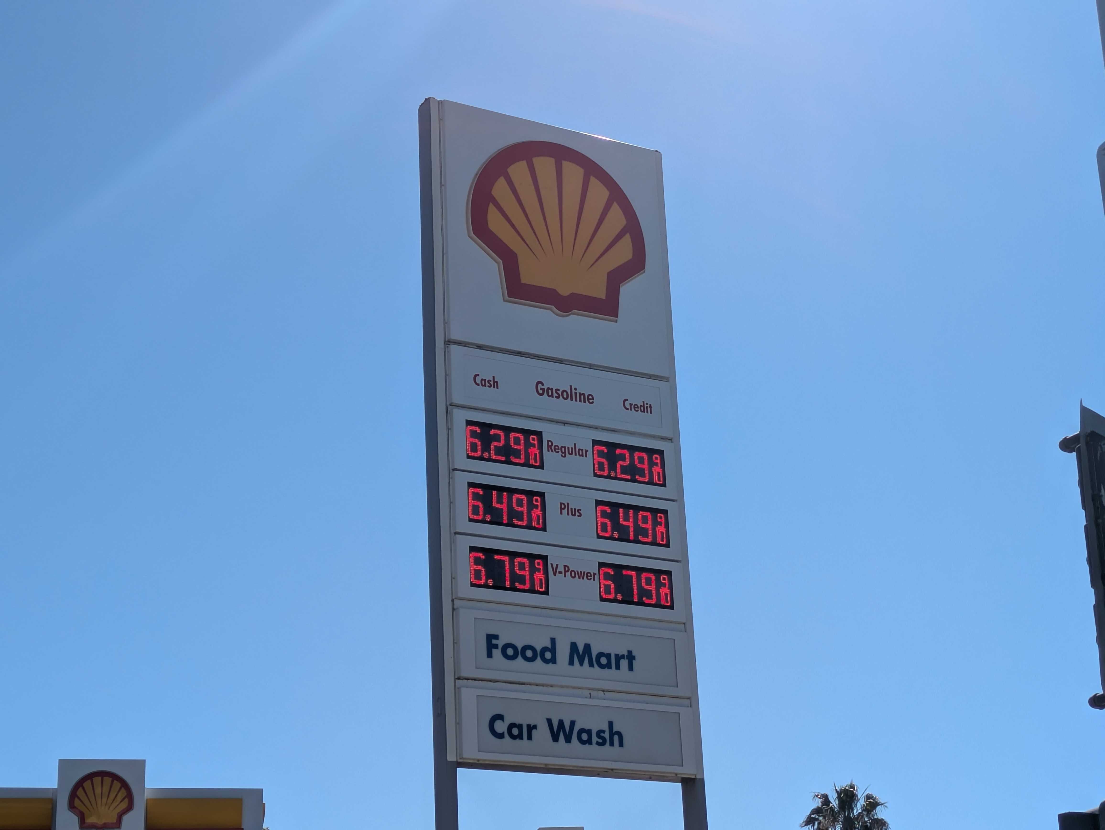 Tall Shell gas station sign against a clear blue sky, featuring the yellow shell logo, a digital price board showing fuel around $6.29–$6.49, and lower panels reading Food Mart and Car Wash.