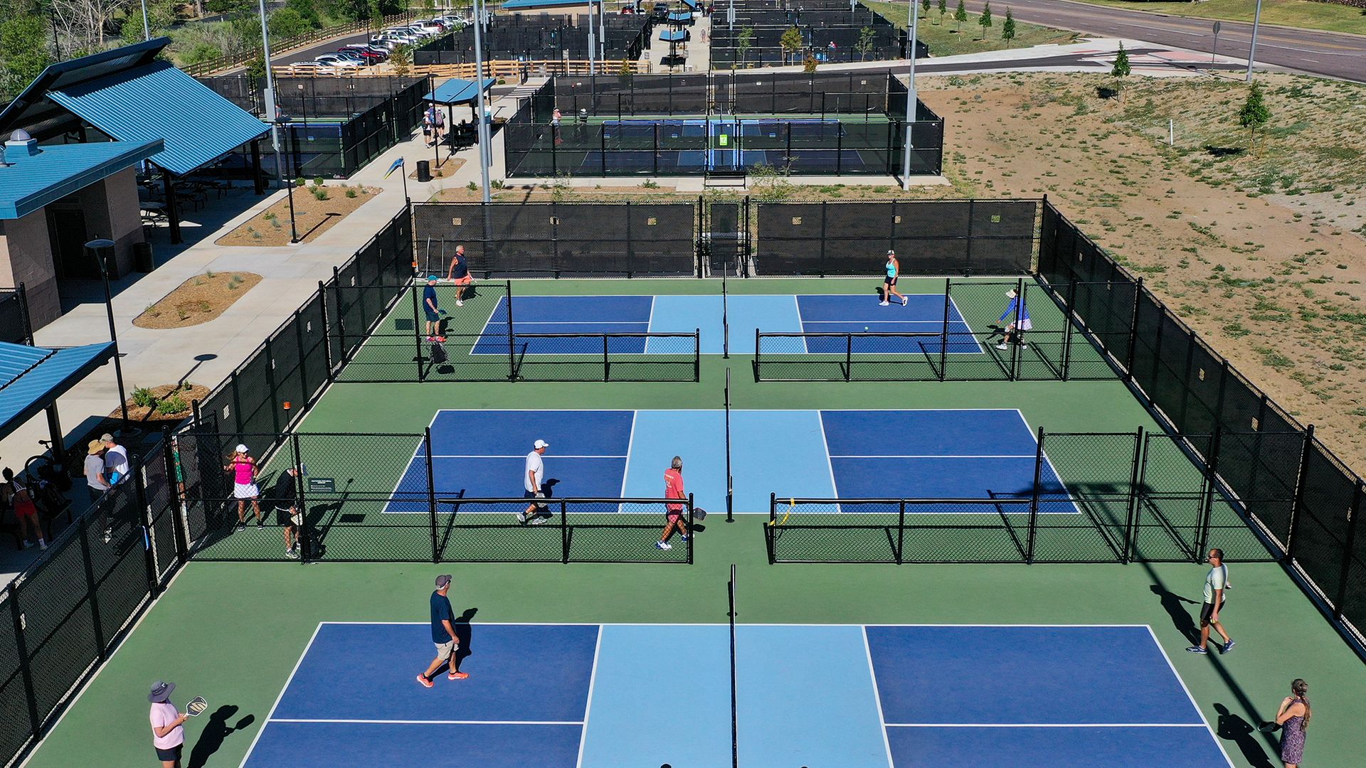 Aerial view of an outdoor sports complex with several fenced blue and light-blue tennis/pickleball courts. Players are on the courts; a blue-roofed building sits on the left, with cars in the background.
