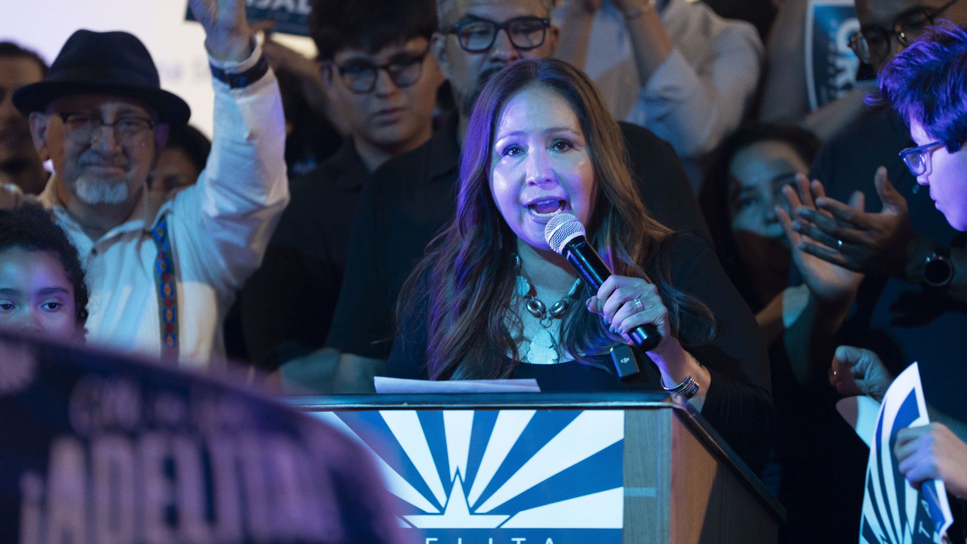 Adelita Grijalva holding a microphone is speaking in front of a podium at a campaign event with a crowd behind her 