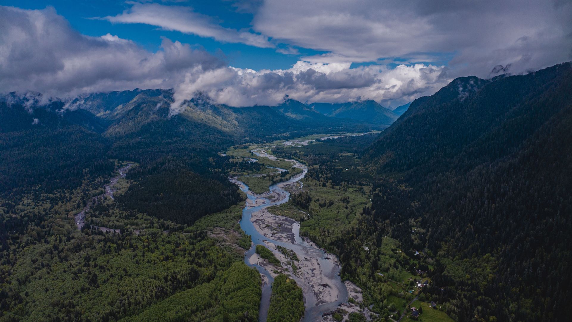A river runs between mountains with puffy clouds in the sky. 