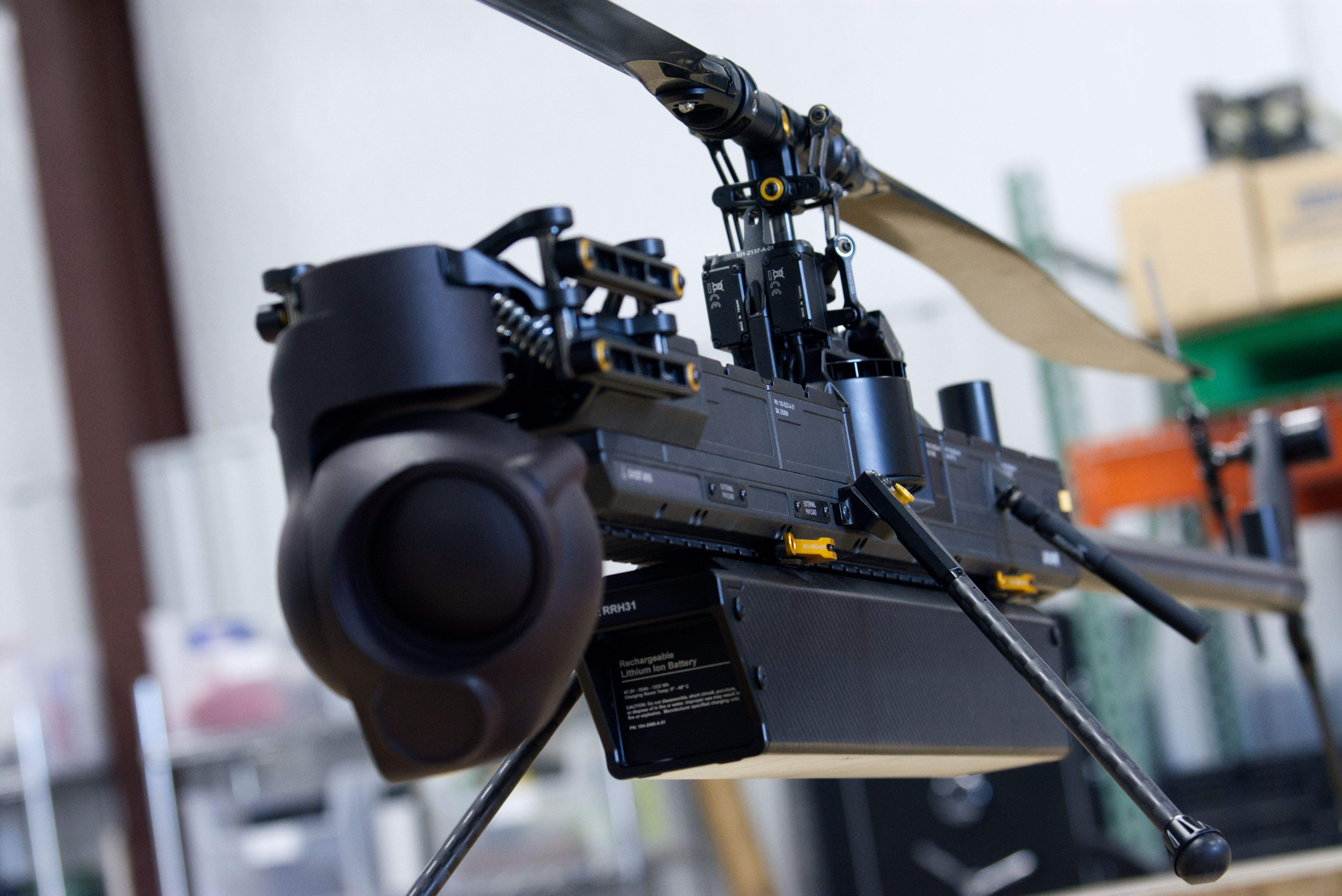 An Anduril drone sits on a table in a workshop. It's black, with large rotor blades and a bulging sensor on the nose.
