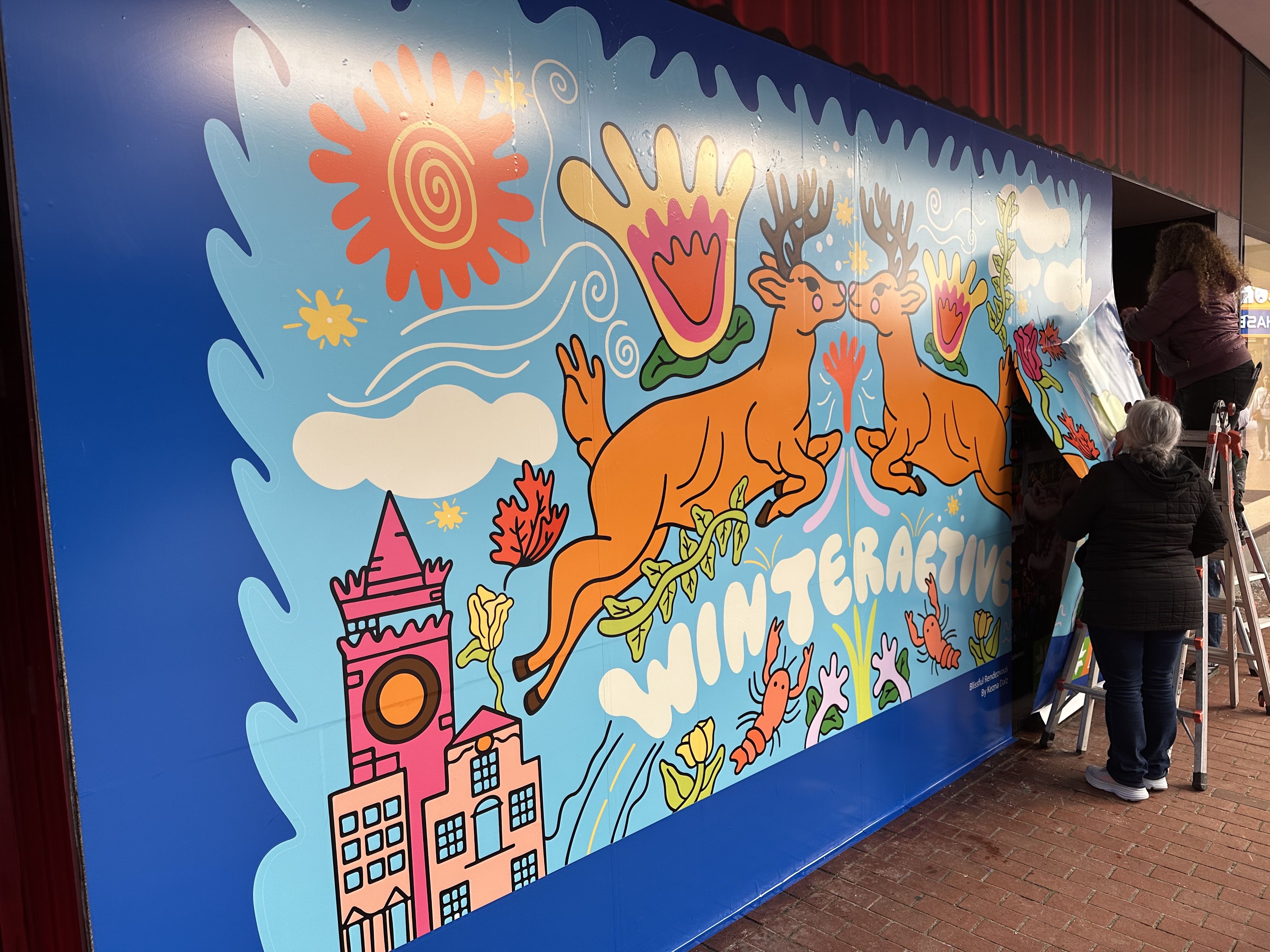 One person on a ladder and another on the ground set up and seal pieces of a colorful mural that says "winteractive" and shows two deer kissing