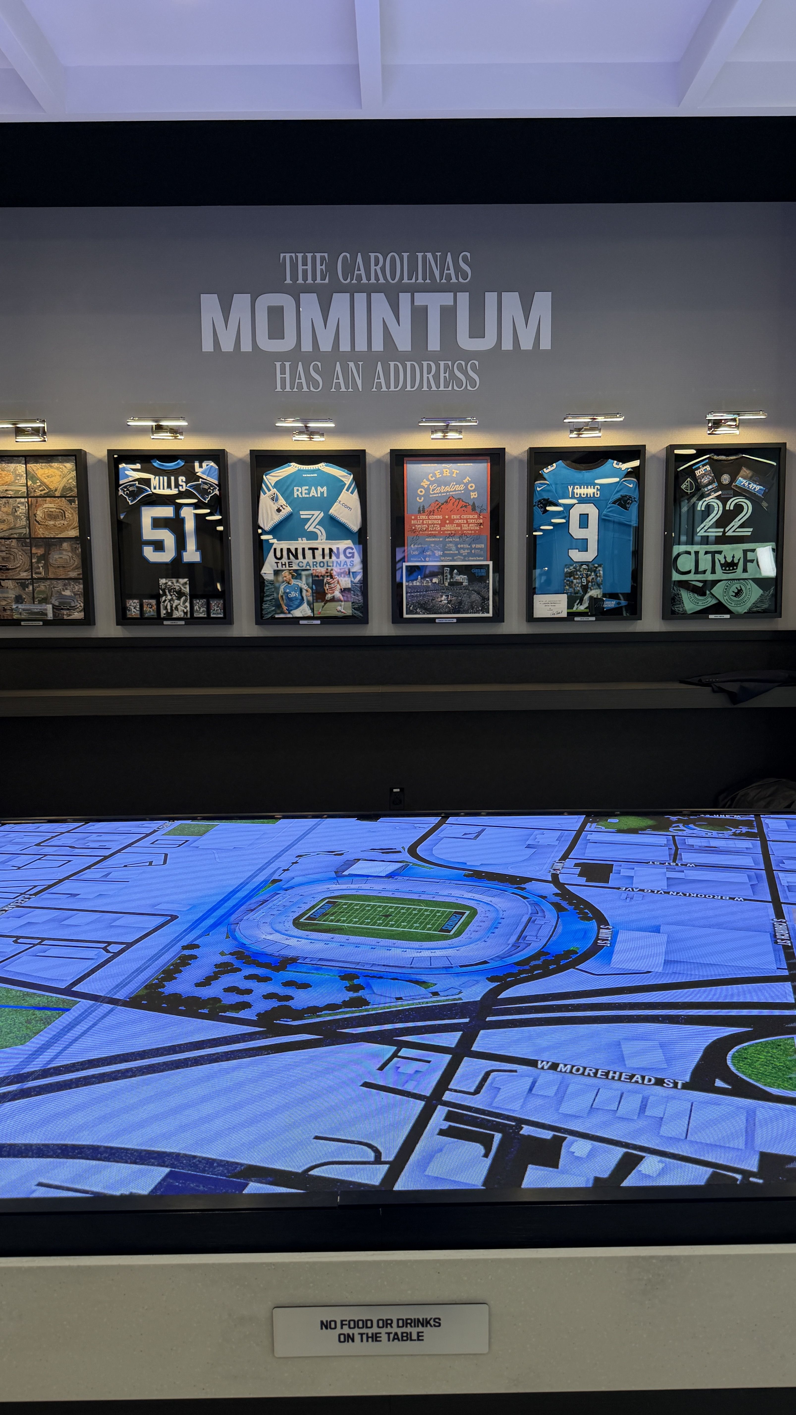Exhibit wall with framed blue Carolina jerseys and posters under large text reading "THE CAROLINAS MOMINTUM HAS AN ADDRESS." A bright blue interactive stadium map table dominates the foreground.