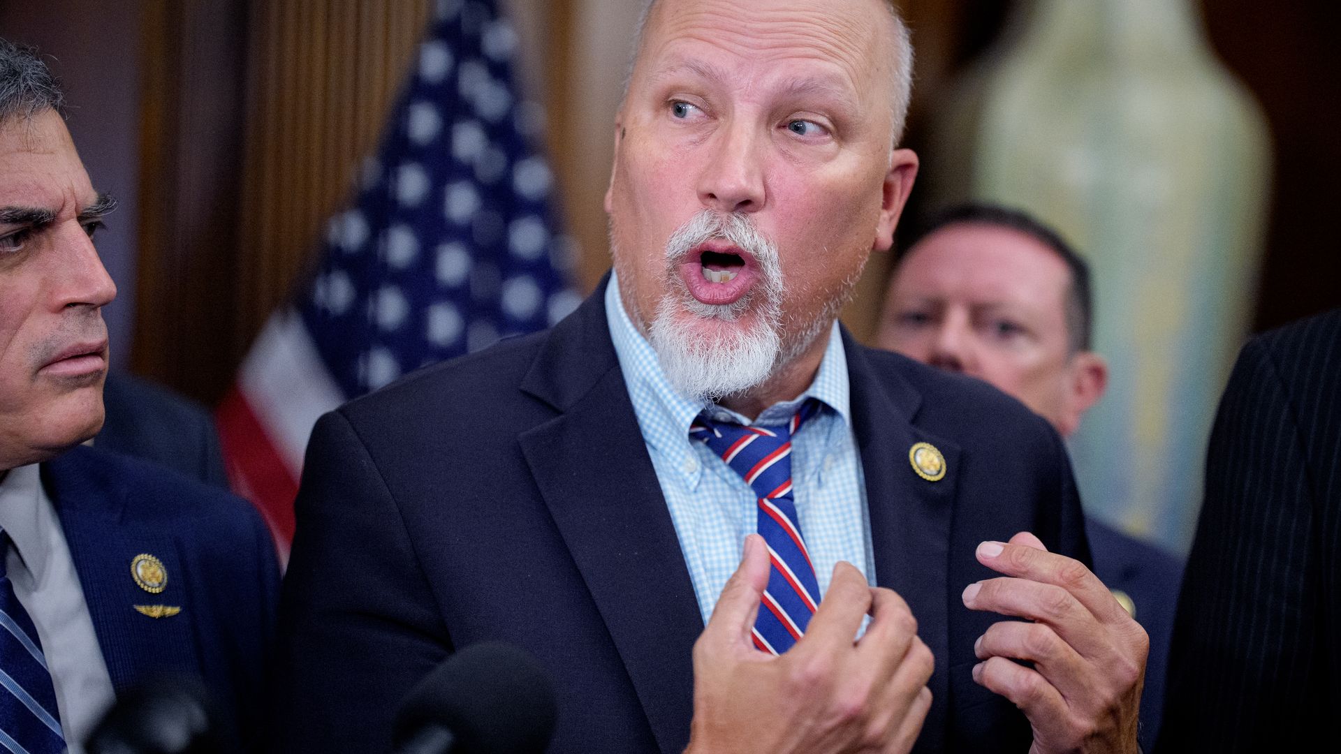 Rep. Chip Roy, flanked by Reps. Andrew Clyde and Eric Burlison, speaks into microphones with his hands raised to chest-level in front of an American flag and a large vase. 