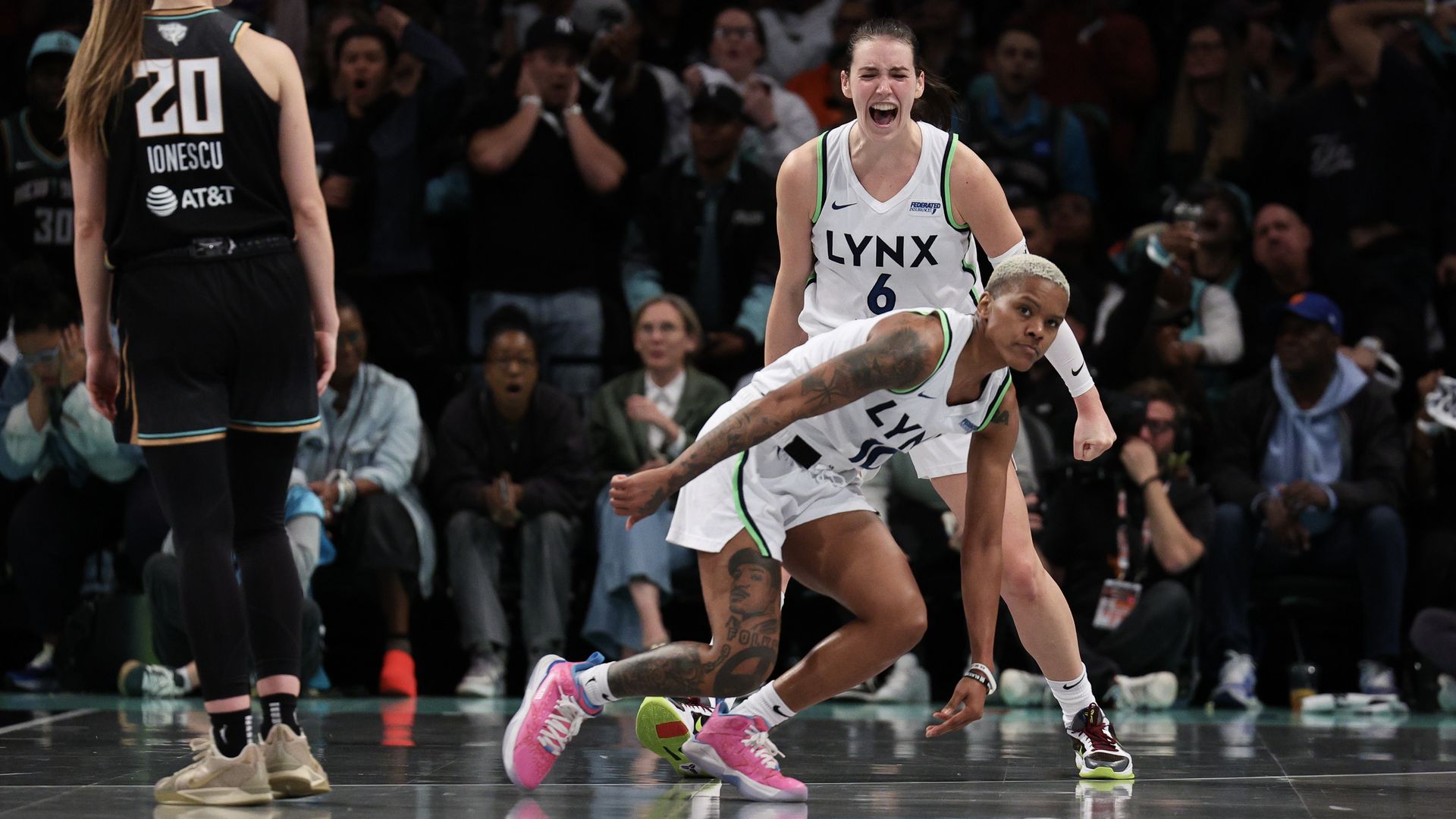 A woman with light skin in a white basketball jersey yells while a teammate with dark skin and bleach-blonde, close-cut hair, falls to the court.