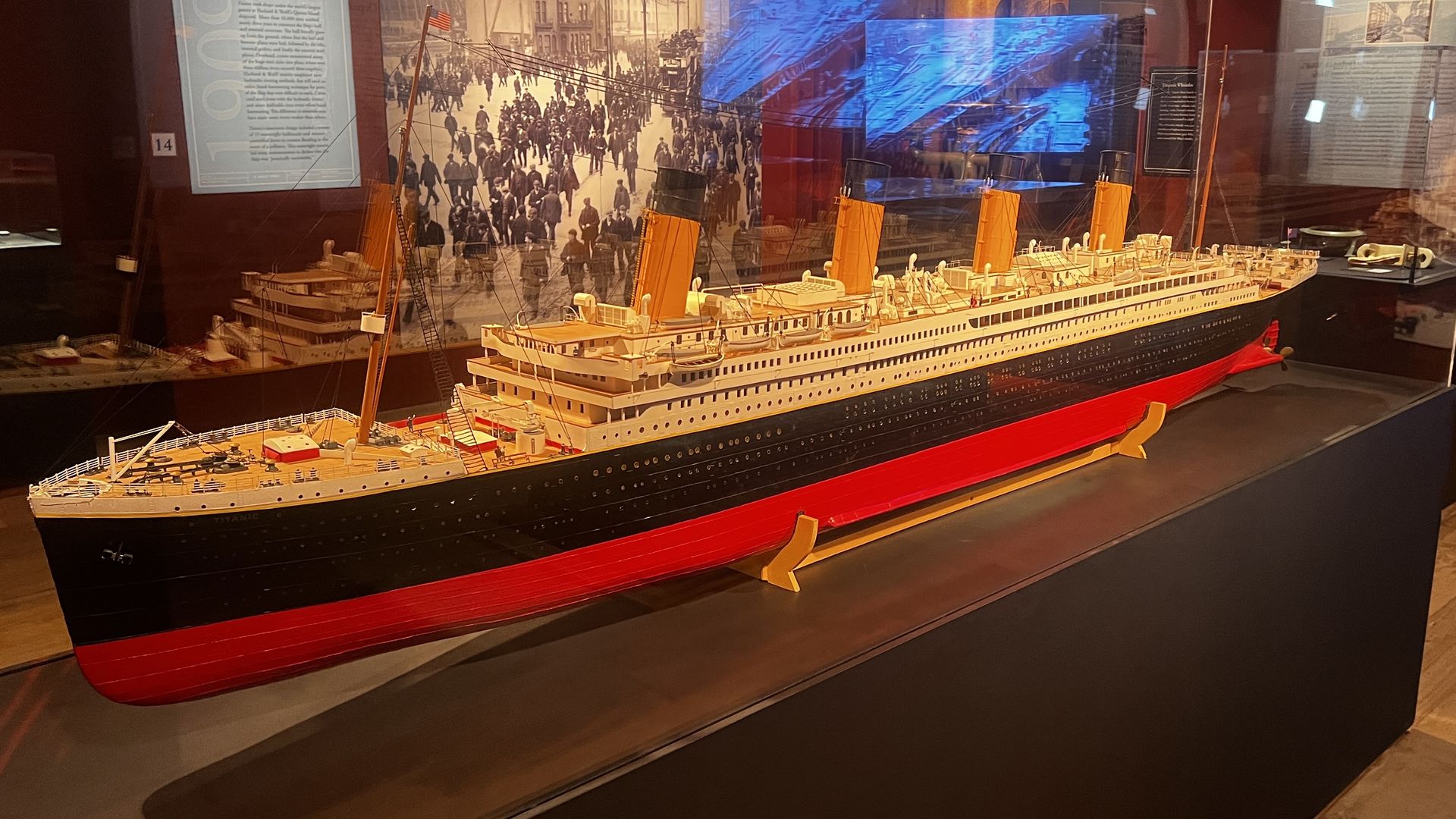 Scale model of the RMS Titanic on display in a museum case; white hull with black lower section and red keel, four yellow funnels, masts and rigging, behind a historical crowd photo.