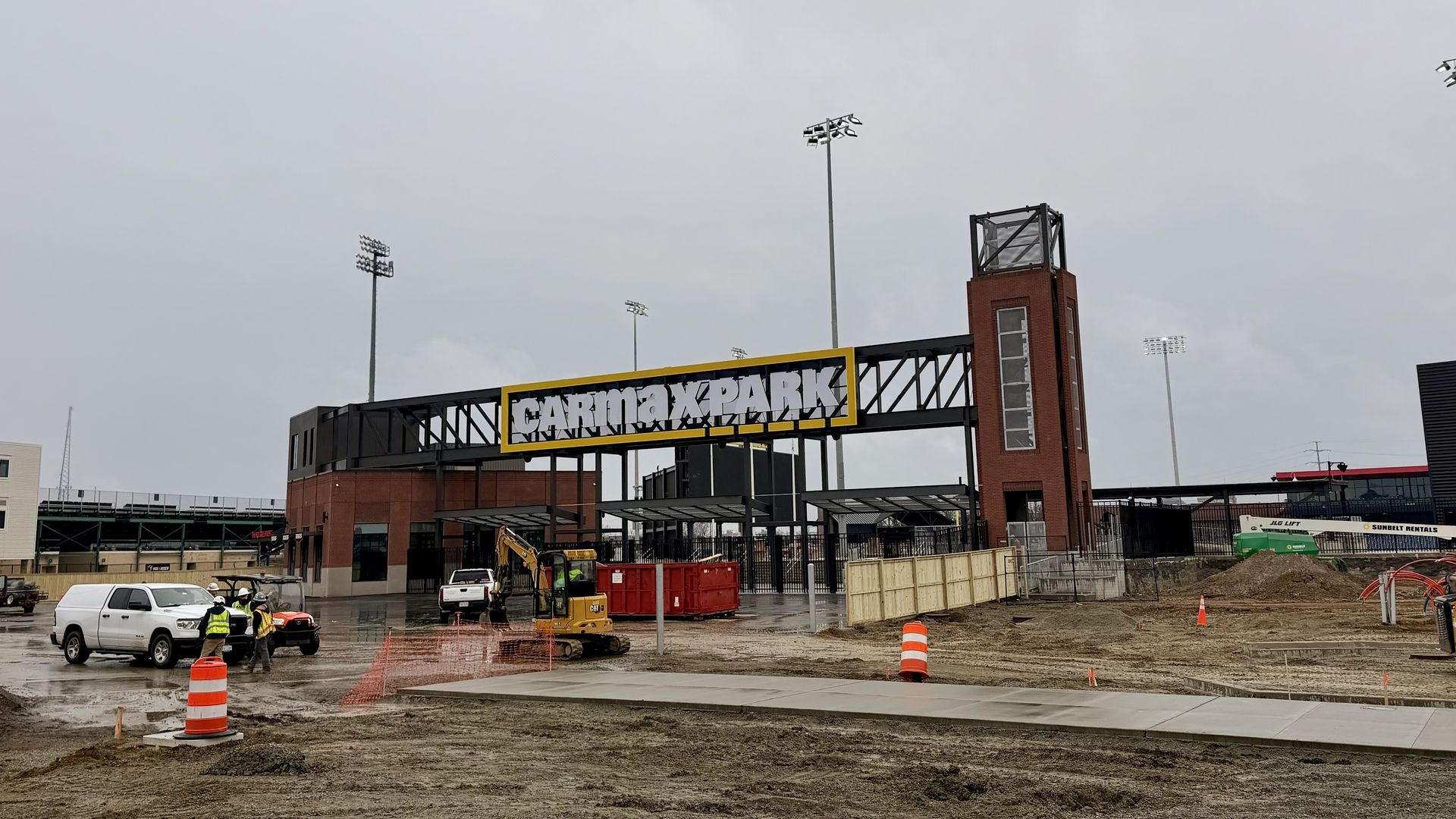 Construction workers and equipment at CarMax Park entrance on a cloudy day, with dirt, orange cones, vehicles, and a brick tower visible.