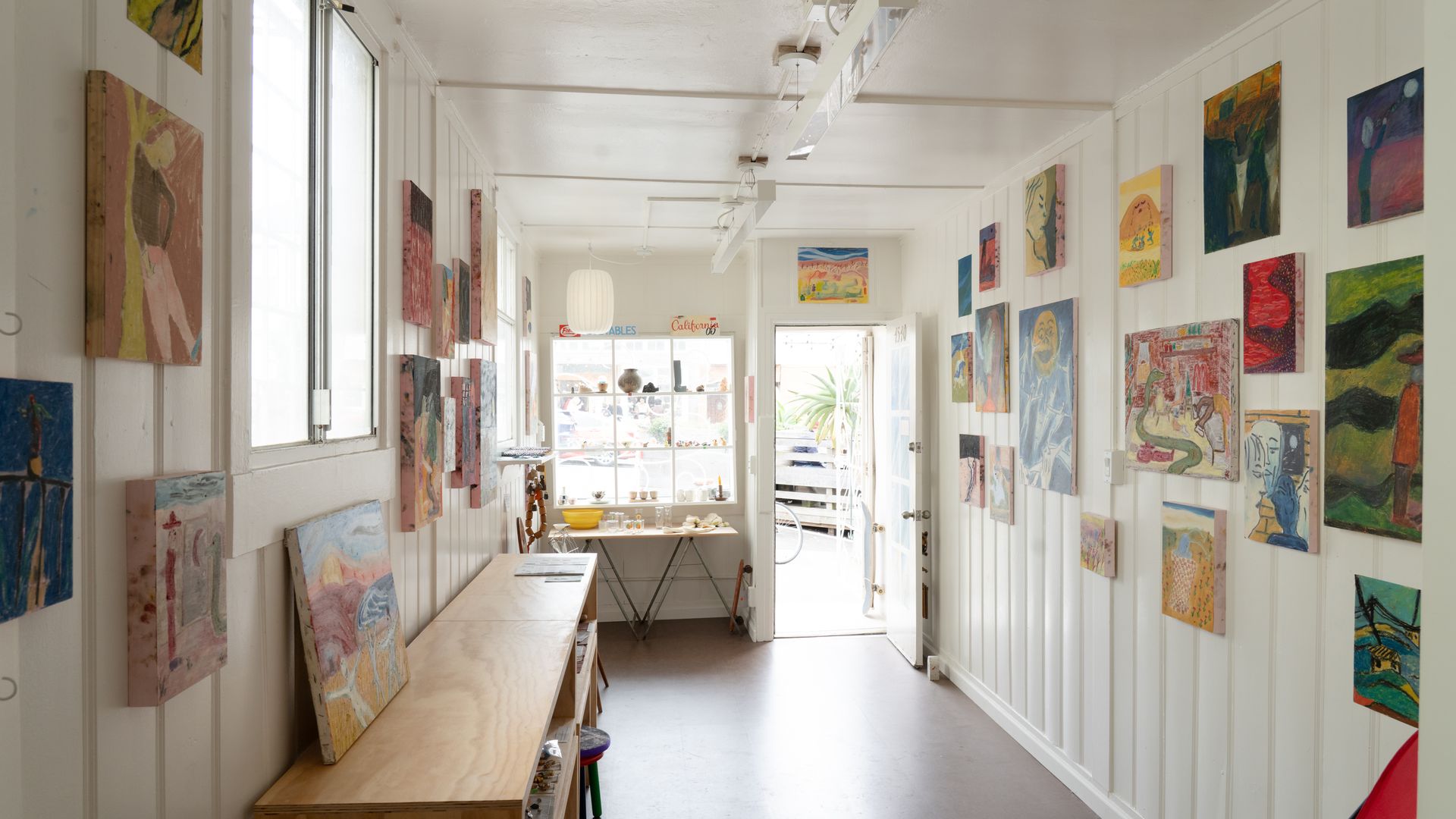 Bright narrow art gallery with white walls, colorful paintings on both sides, large window at the end, wooden shelves, a table with items, and an open door letting in daylight.