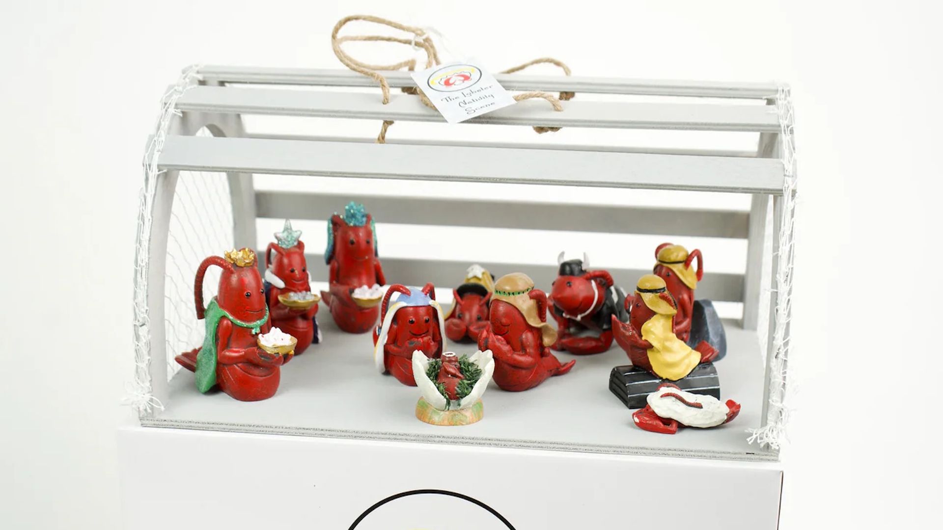 Miniature red lobster figurines arranged in a nativity scene inside a white lobster trap with a label reading "Lobster Nativity Scene" on a white box below.