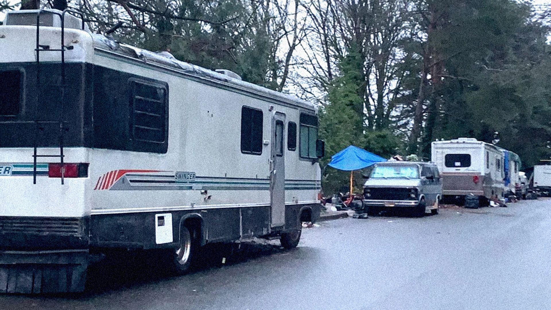 Several parked RVs and a blue tarp shelter along a tree-lined street, with scattered belongings and a van among the vehicles under overcast sky.