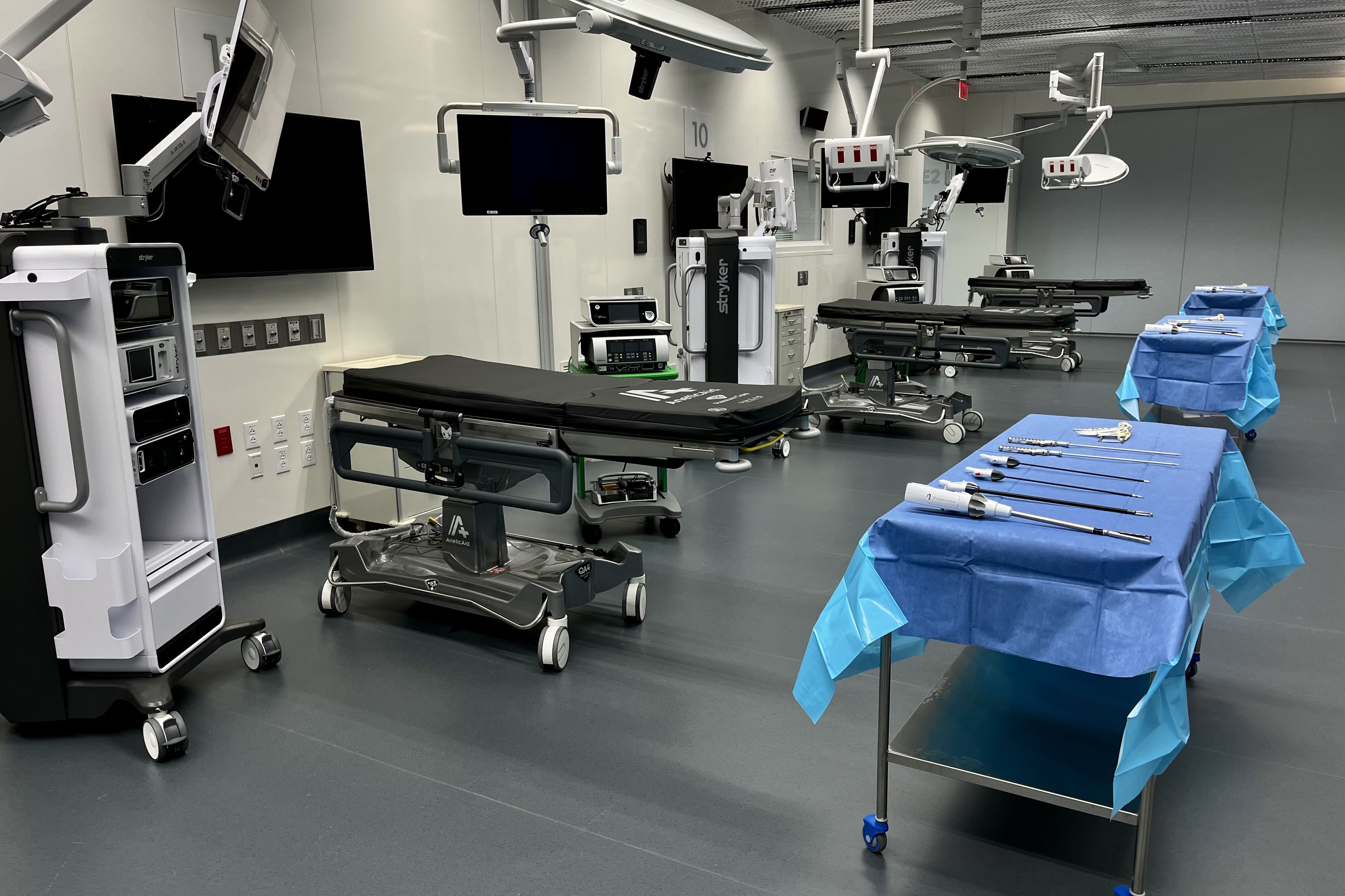 Modern medical operating room with three black patient beds, multiple screens and surgical lights, and instrument tables covered in blue sterile drapes holding surgical tools.