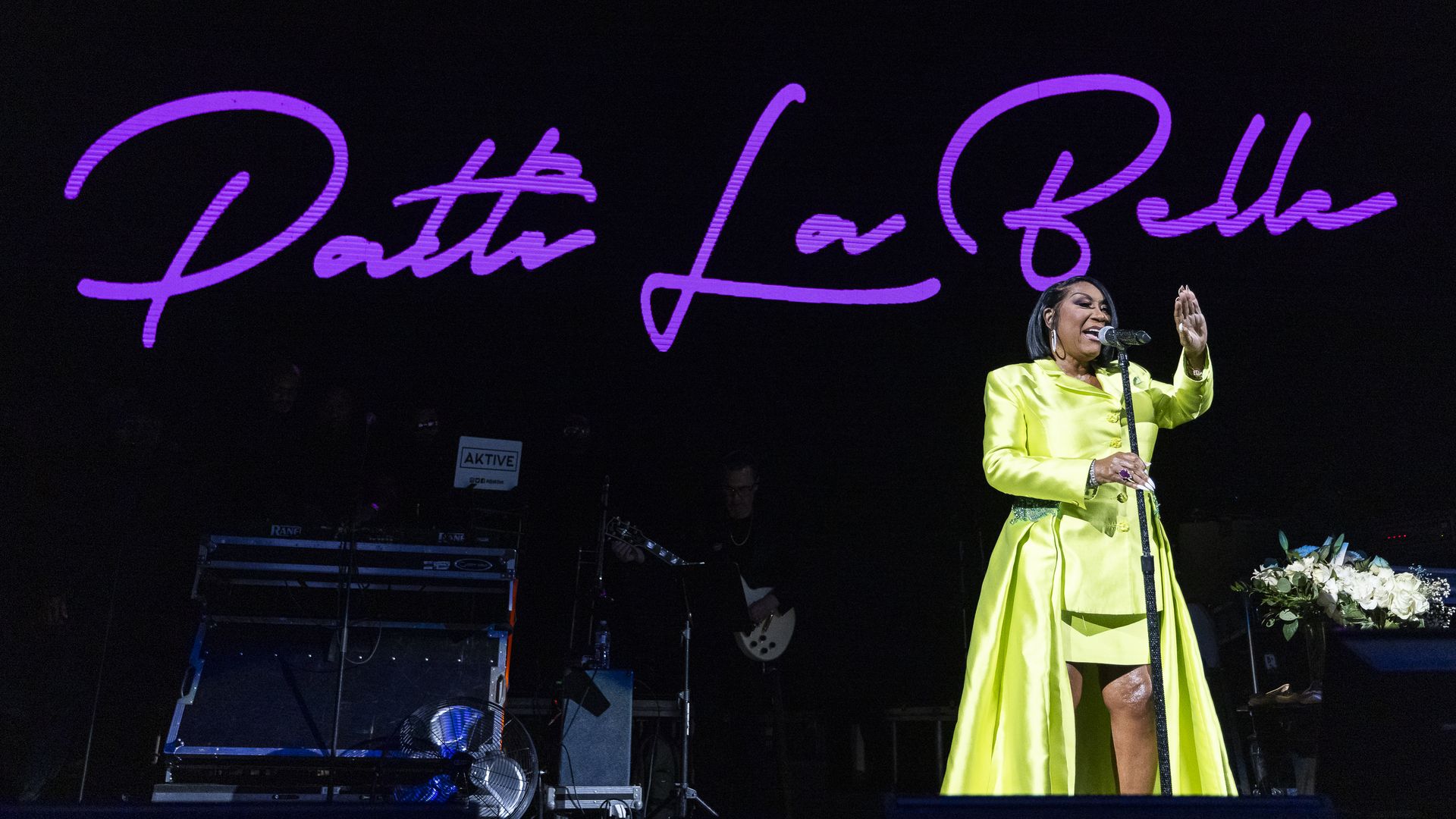 Patti LaBelle performs on a dark stage in a bright neon-yellow dress, singing into a mic as a large purple cursive "Patti LaBelle" sign glows behind her; stage gear and flowers nearby.