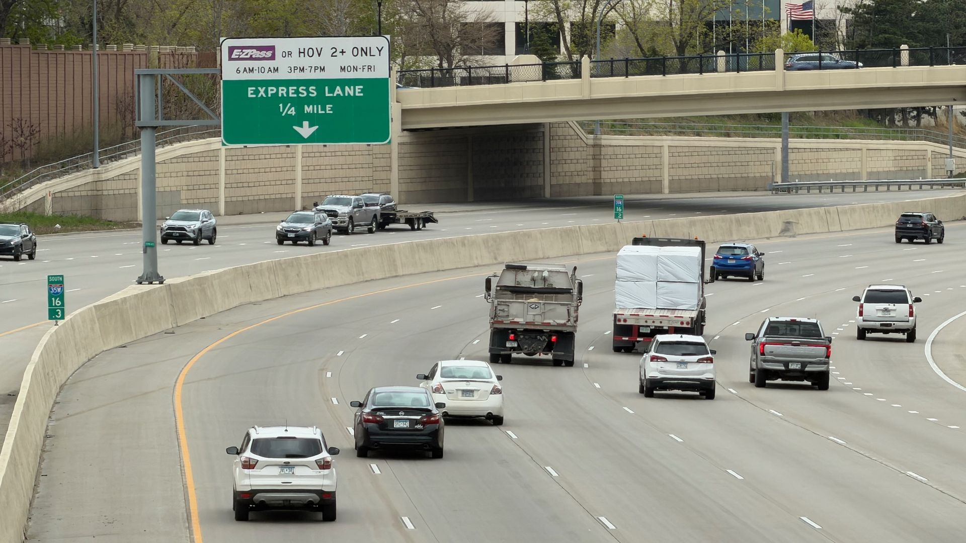 Multi-lane highway beneath an overpass, with a green sign reading "EXPRESS LANE 1/4 MILE" and a downward arrow. Traffic includes cars and a trailer; background shows office buildings, trees, and an American flag.