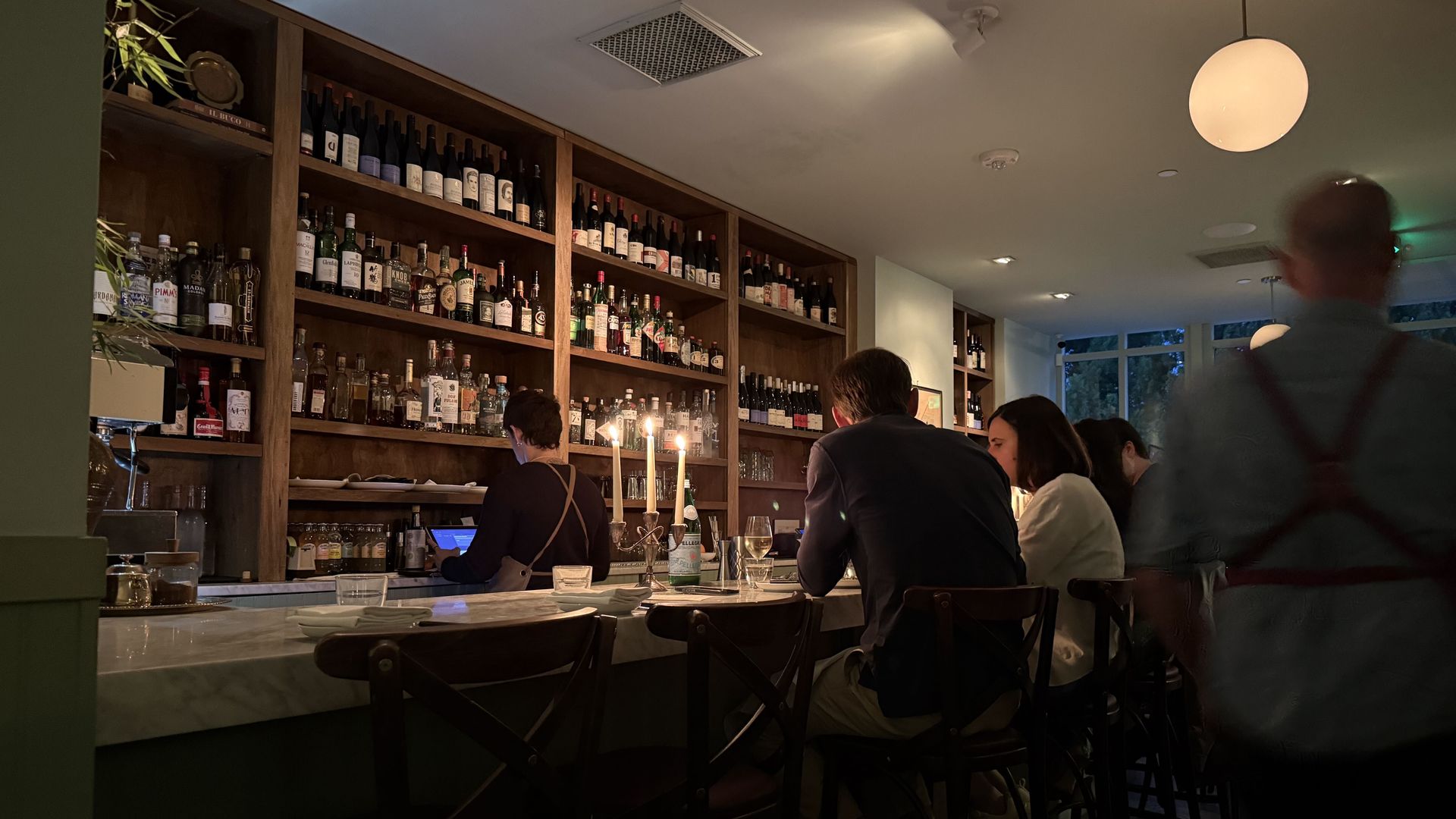 Inside a cozy bar with wooden shelves lined with bottles behind the counter. Warm lighting, candles on the bar, patrons seated at stools, a round pendant lamp and dim ceiling lights.