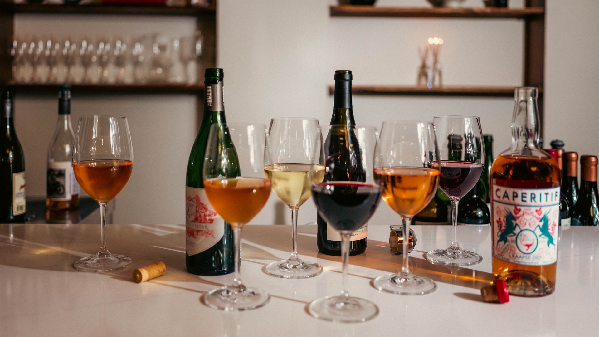 Several wine glasses filled with amber, white, and red wine stand on a white surface with wine bottles, including one labeled "CAPERITIF," and corks scattered around.