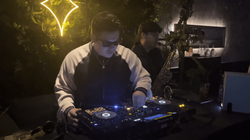 A DJ adjusts his turntable next to a man playing the saxophone at a dark bar. 