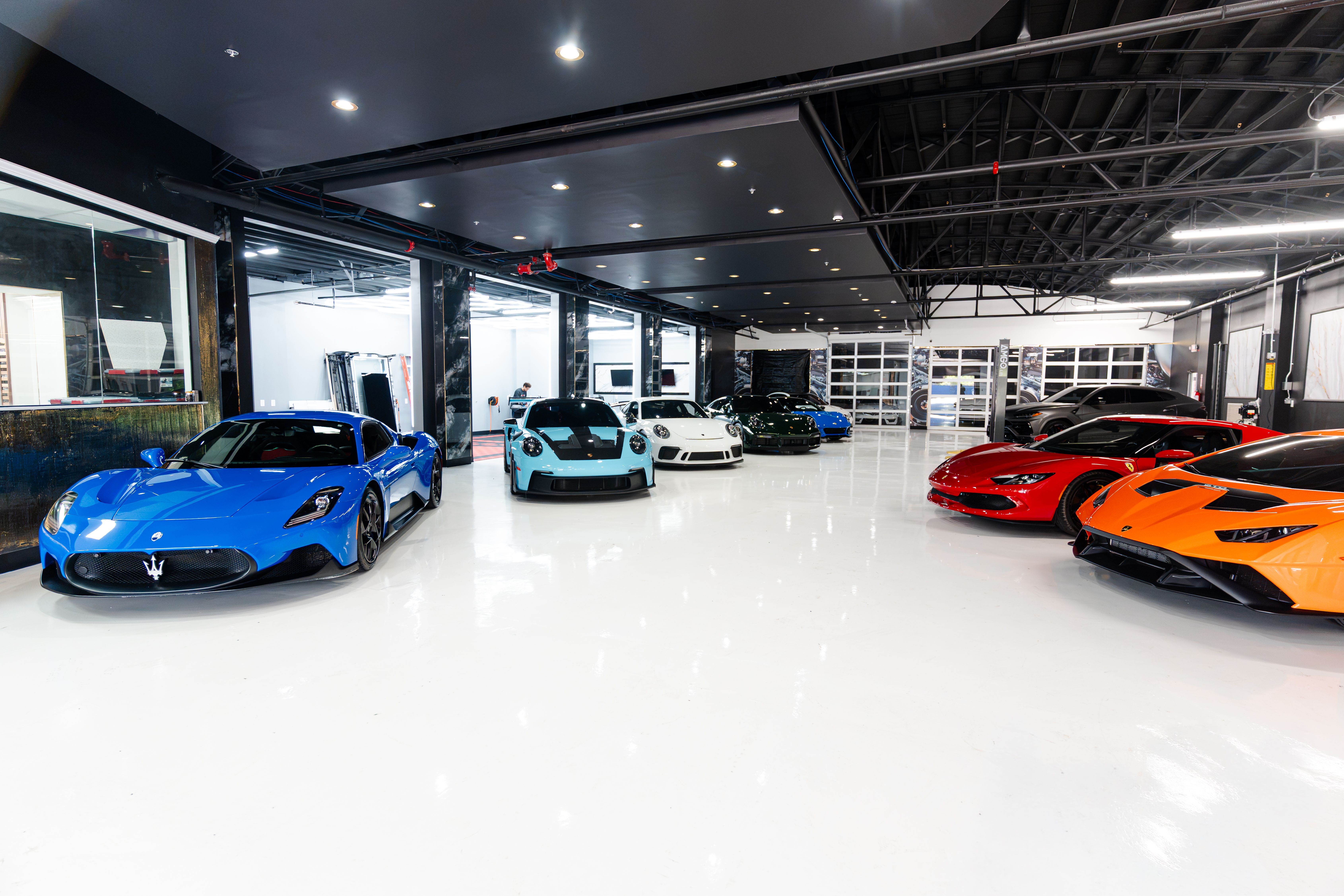 A modern car showroom showcasing a lineup of colorful sports cars (blue, teal, white, green, red, orange) on a glossy white floor under an exposed metal ceiling with bright lights.