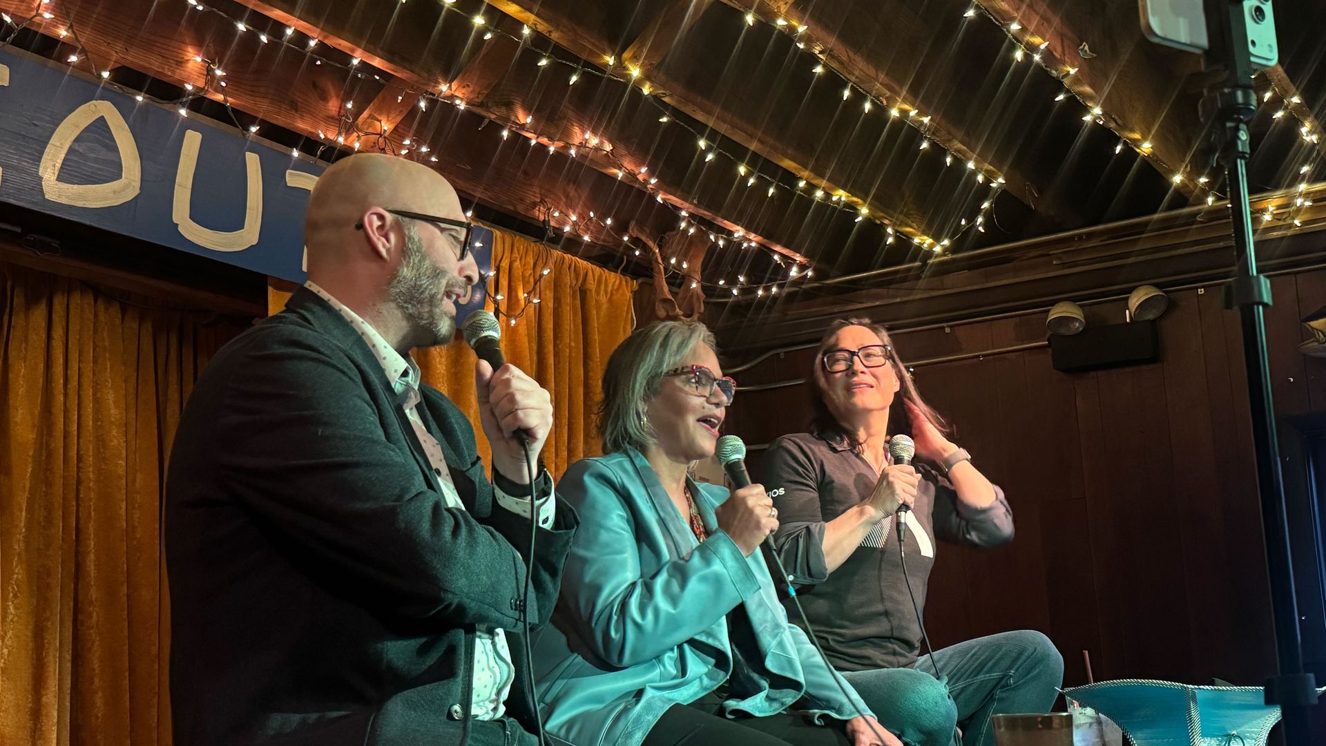 Three panelists with microphones on a stage: a bald man in a blazer, a woman in teal, and a woman in gray. Warm string lights, amber curtains, a banner with "OU" visible, and a smartphone on a tripod.