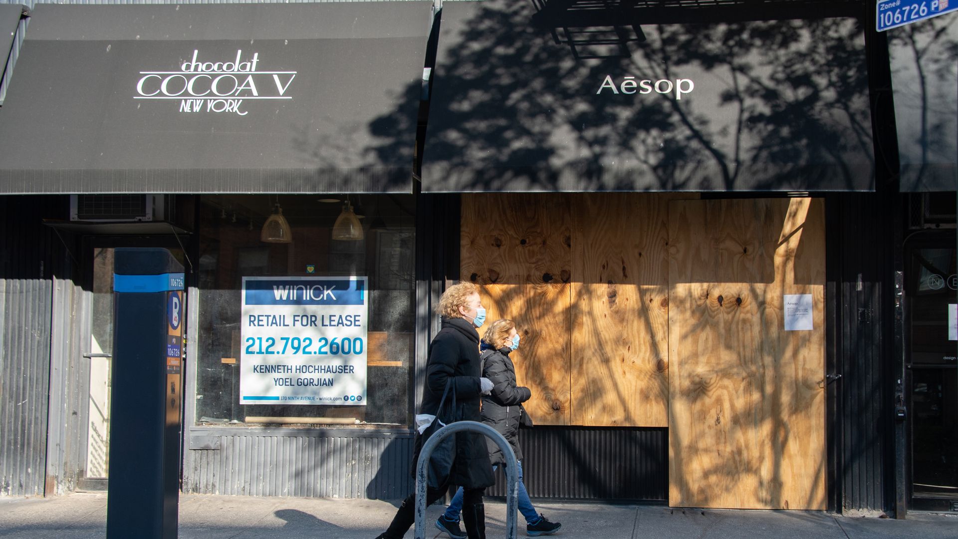 Two women wearing protective masks and gloves walk past a retail space for lease and a boarded up shop