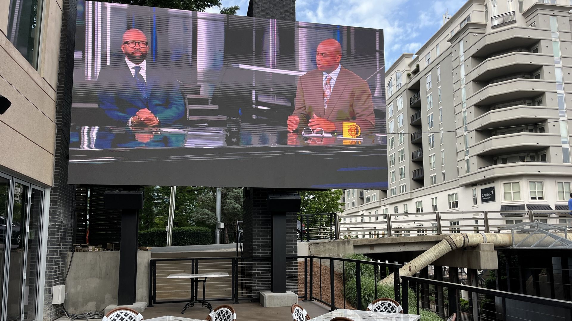 Outdoor plaza with a large LED screen showing two men in suits at a desk. Foreground features white marble tables and black-and-white patterned chairs; modern buildings and a blue sky behind.
