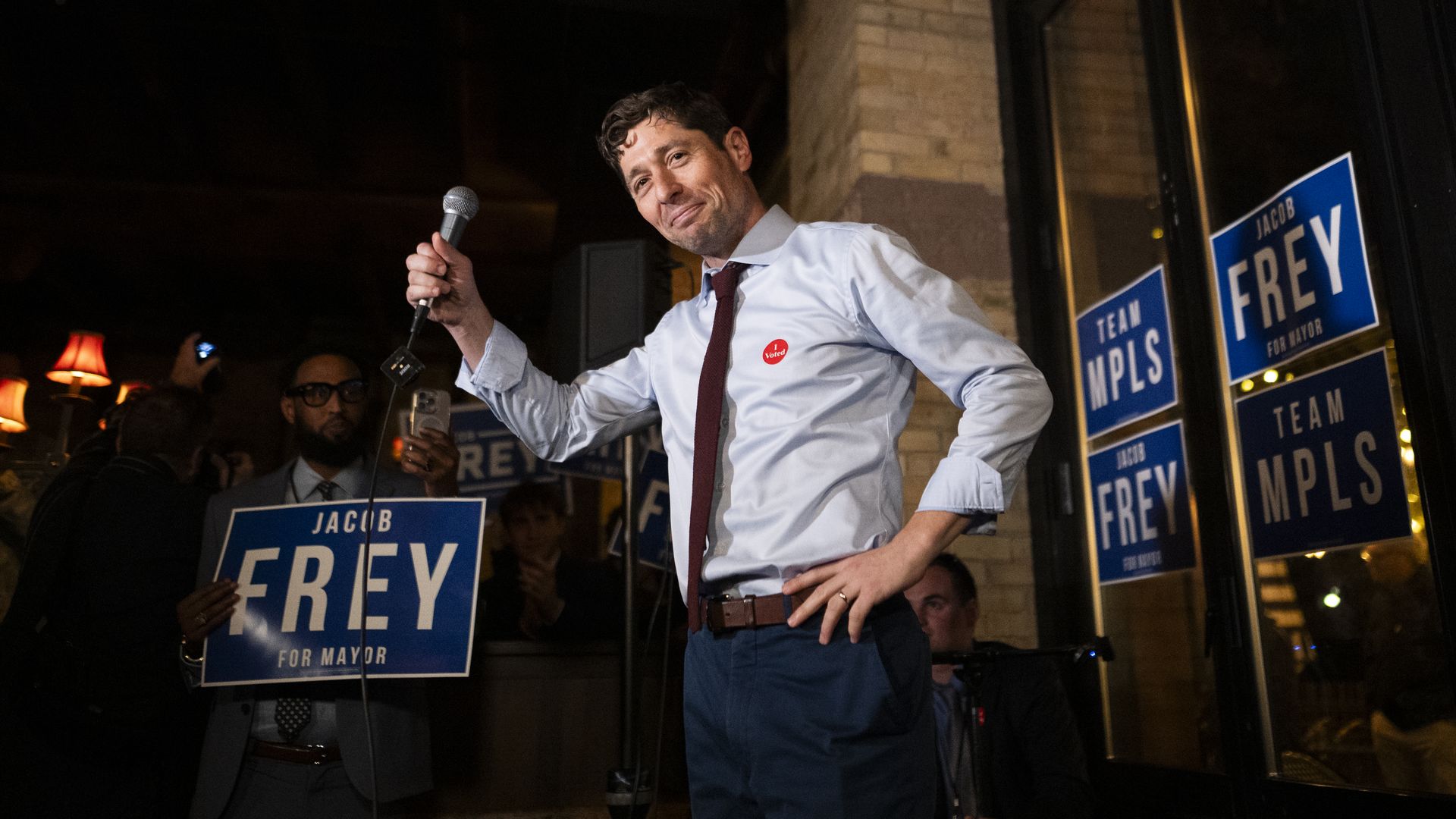 Mayor Jacob Frey wearing a dress shirt and tie, holds a microphone in front of campaign signs. 