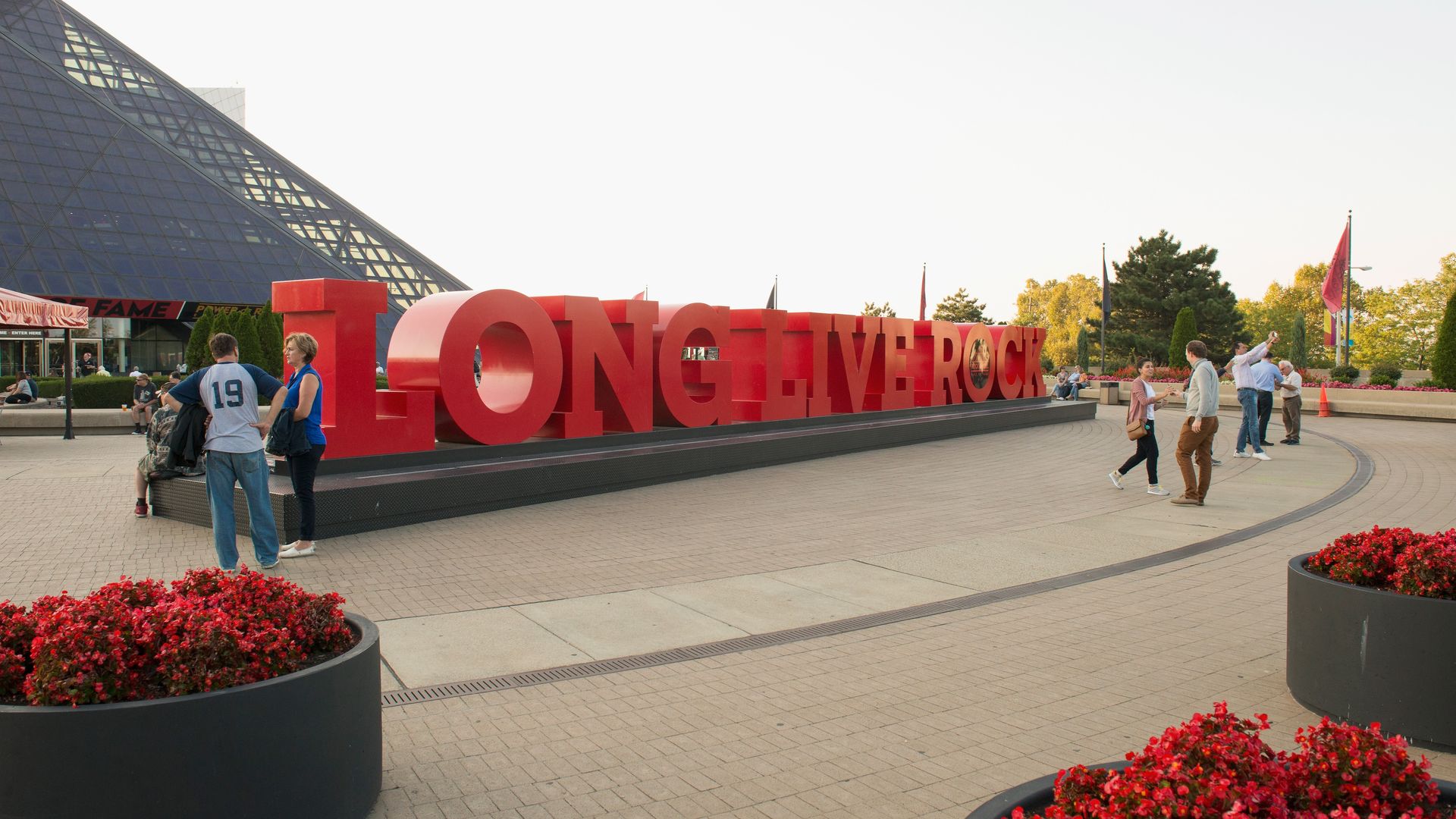 A large red sculpture spelling "LONG LIVE ROCK" sits on a black pedestal outside a glass pyramid building. People chat and walk nearby, with red flower planters in the foreground.
