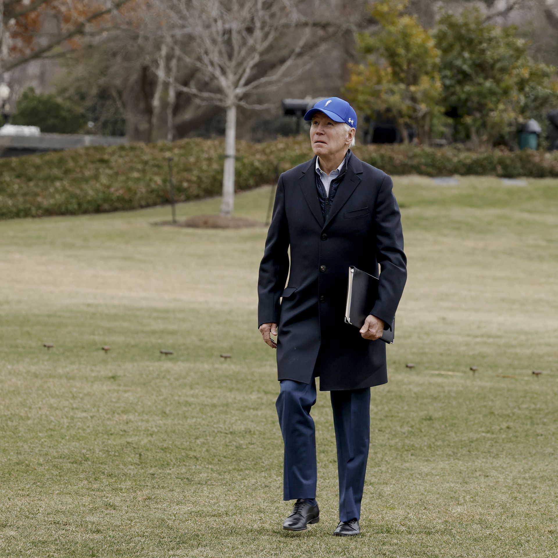 Joe Biden walks in winter grass on the White House south lawn, looking up the sky in his blue baseball cap