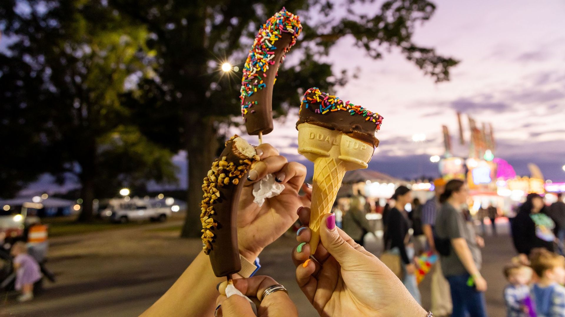 Several hands hold up chocolate-dipped ice creams with rainbow sprinkles at a twilight carnival, with a purple sky and colorful lights in the background.