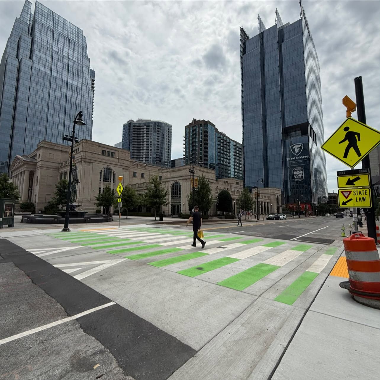 Urban street with tall glass skyscrapers, a beige historic building, and a green striped crosswalk. A person crosses as yellow pedestrian signs and a traffic barrel appear nearby.