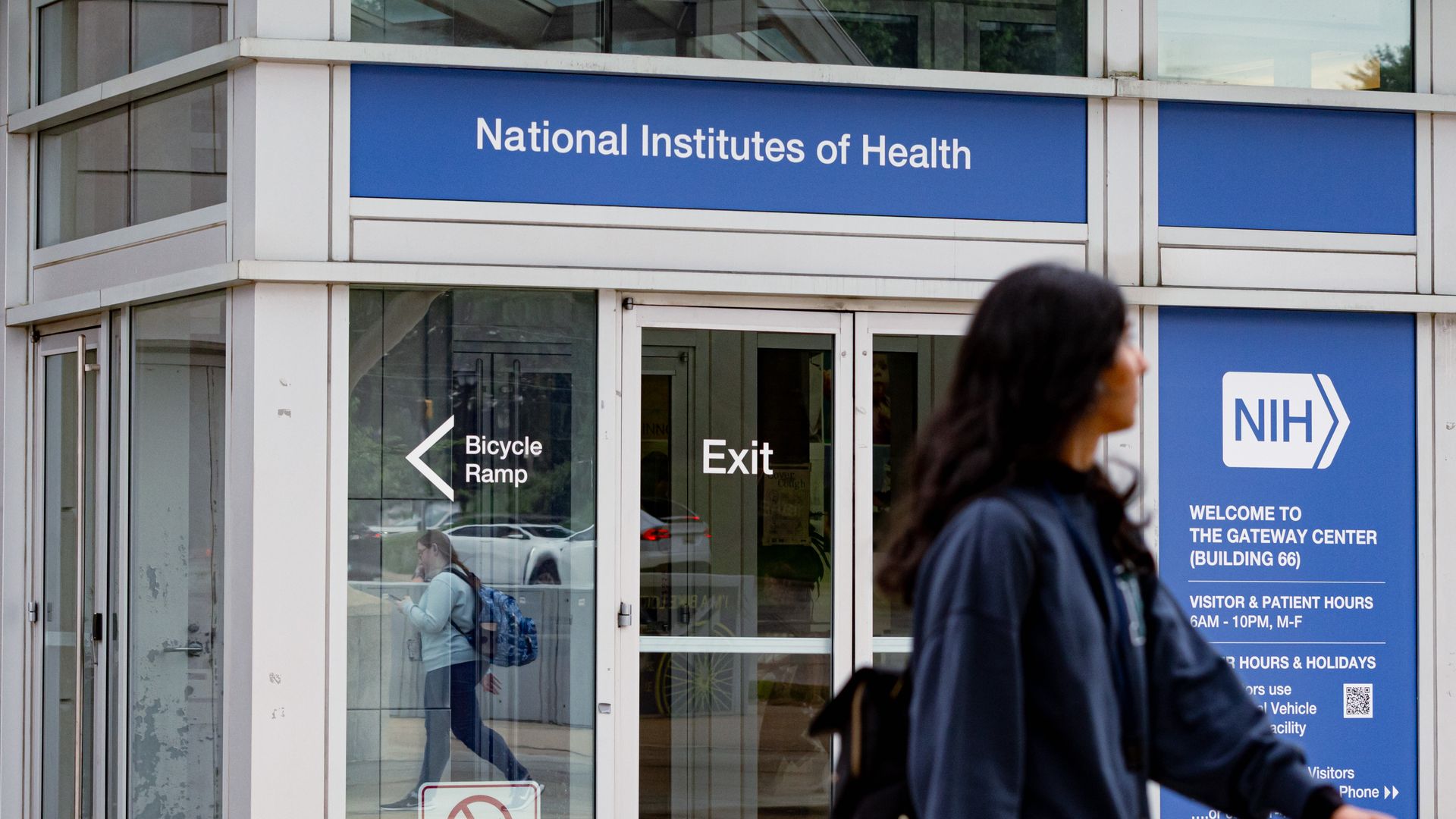 Entrance of the National Institutes of Health Gateway Center with blue signs, glass doors marked Exit and Bicycle Ramp, a no firearms sign, and a person walking past carrying a backpack.